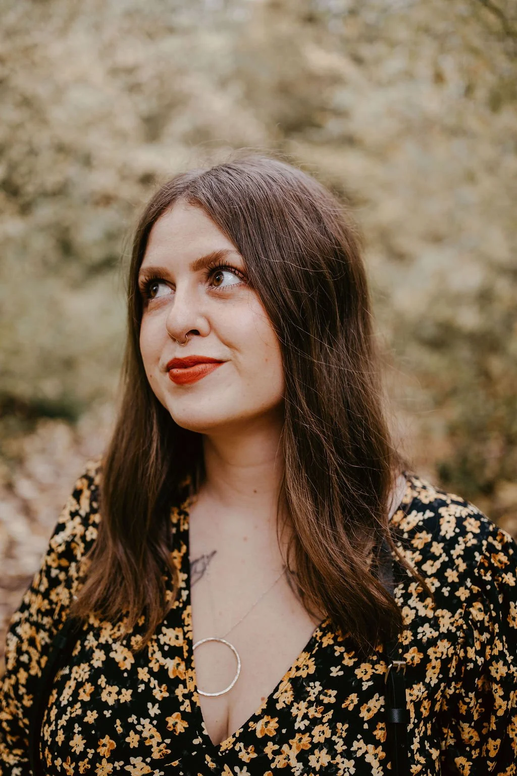 A woman with long brown hair and freckles, wearing a floral black top with a round silver necklace, standing outdoors with a blurred background of trees and leaves.