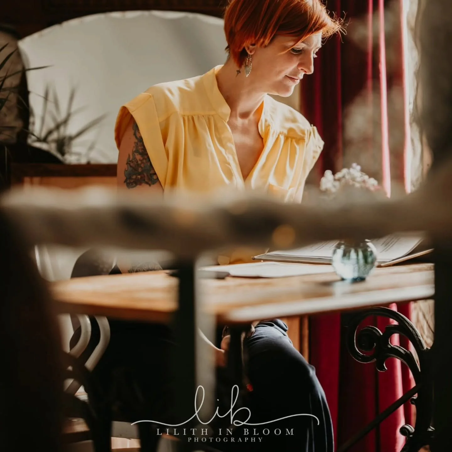 A woman with short red hair wearing a yellow shirt is sitting at a wooden table with an open book and a small glass vase with flowers.