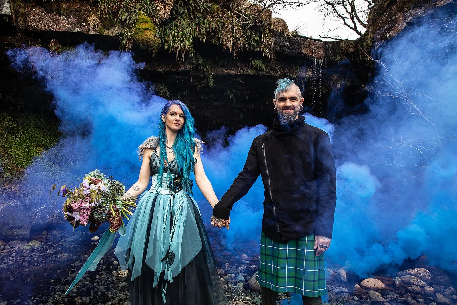A couple holding hands in front of blue smoke with a rocky, green, and mossy background. The woman has long, curly blue hair, tattoos, and a colorful dress, holding a bouquet of flowers. The man has short, blue hair, a beard, tattoos, and is dressed 