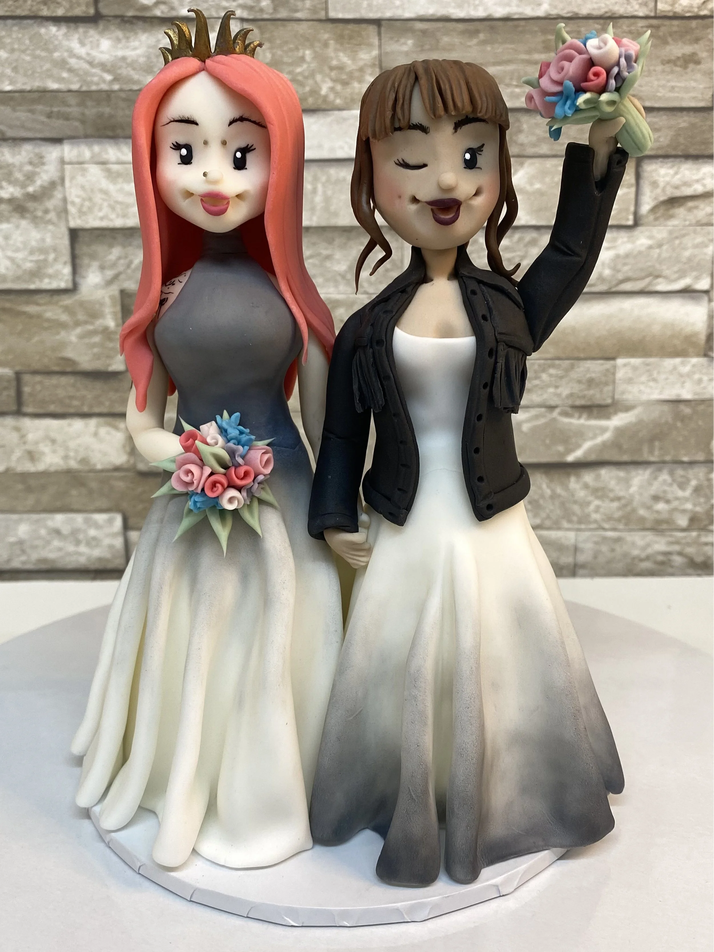 Cake topper figurines of two women in wedding dresses, one holding a bouquet and the other with a flower crown, posing against a stone wall background.