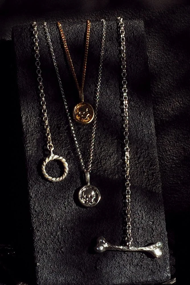 Four necklaces with different pendants displayed on a black surface. One has a circular pendant with a smiley face, another has a similar central face with a different chain, the third has a circular twisted loop, and the fourth features a bone-shape