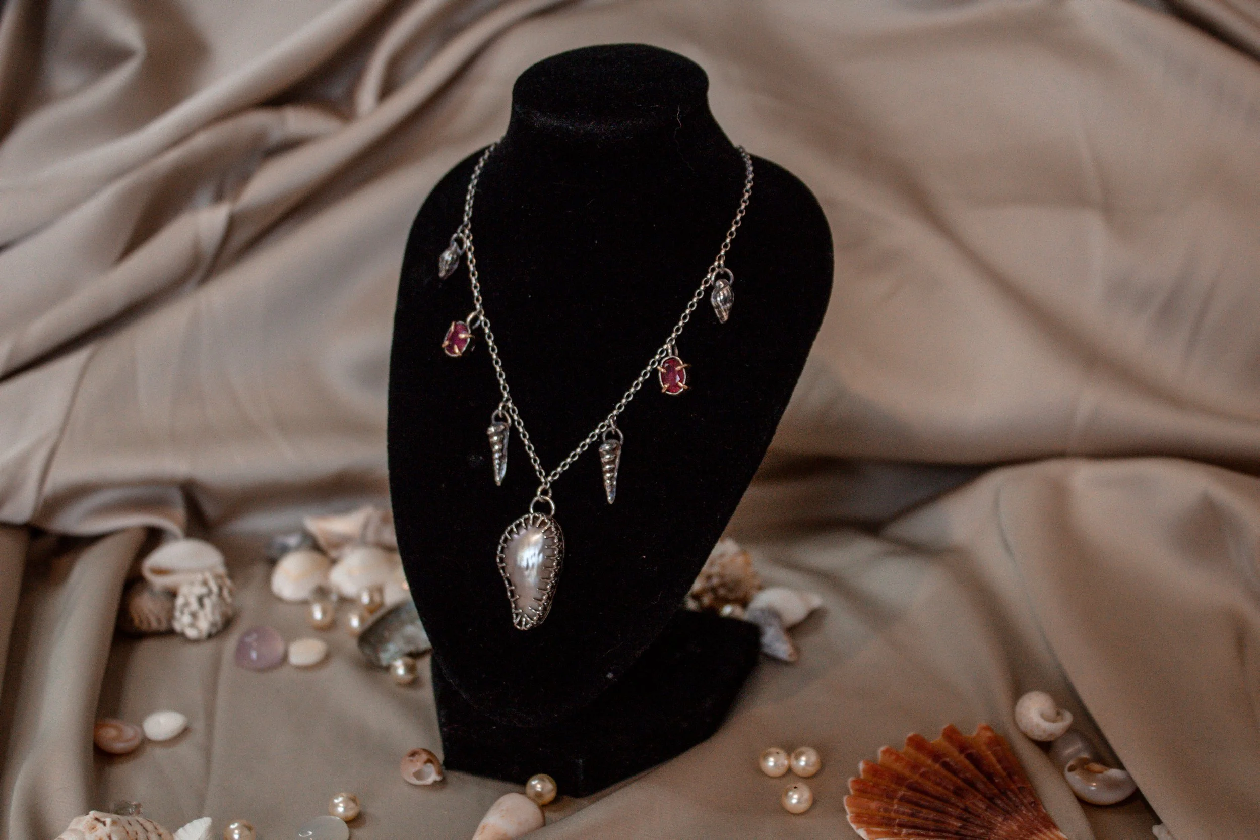 A silver necklace with shell-shaped pendants displayed on a black velvet bust, surrounded by seashells, pearls, and coral on a beige fabric background.