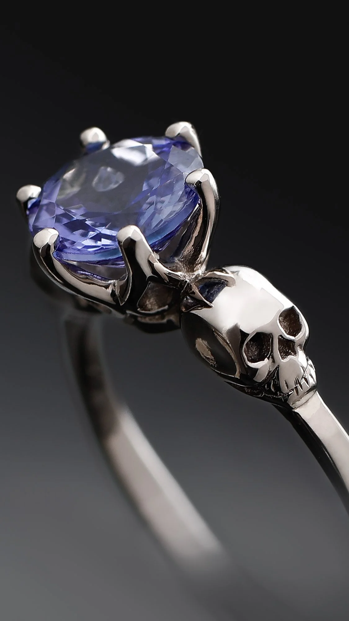 A silver ring with a skull design holding an oval-shaped purple gemstone.