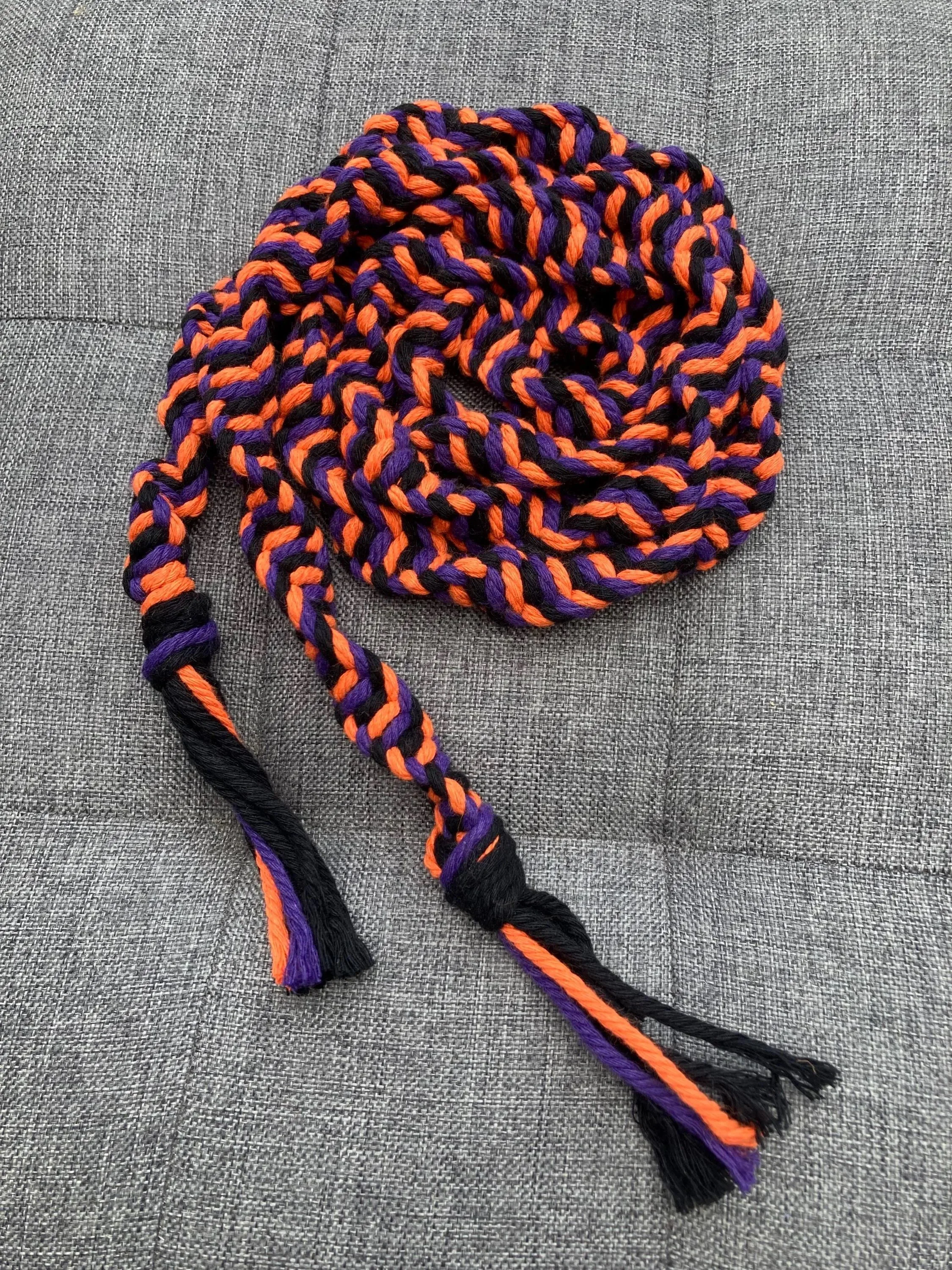 A multicolored woven scarf with black, purple, and orange strands, loosely coiled on a gray fabric surface.