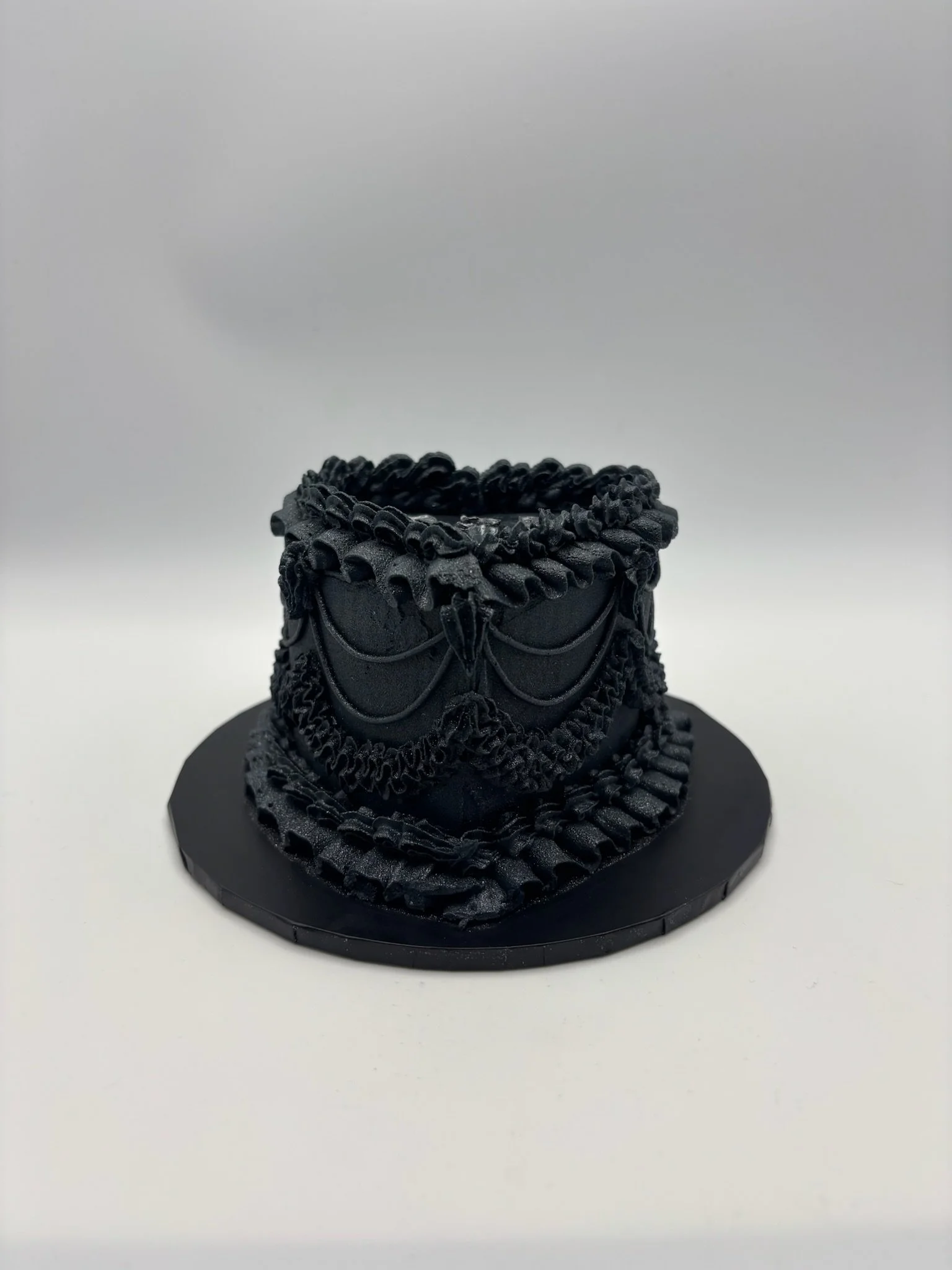 A black decorative mask with intricate ruffles and swag details displayed on a black circular base.