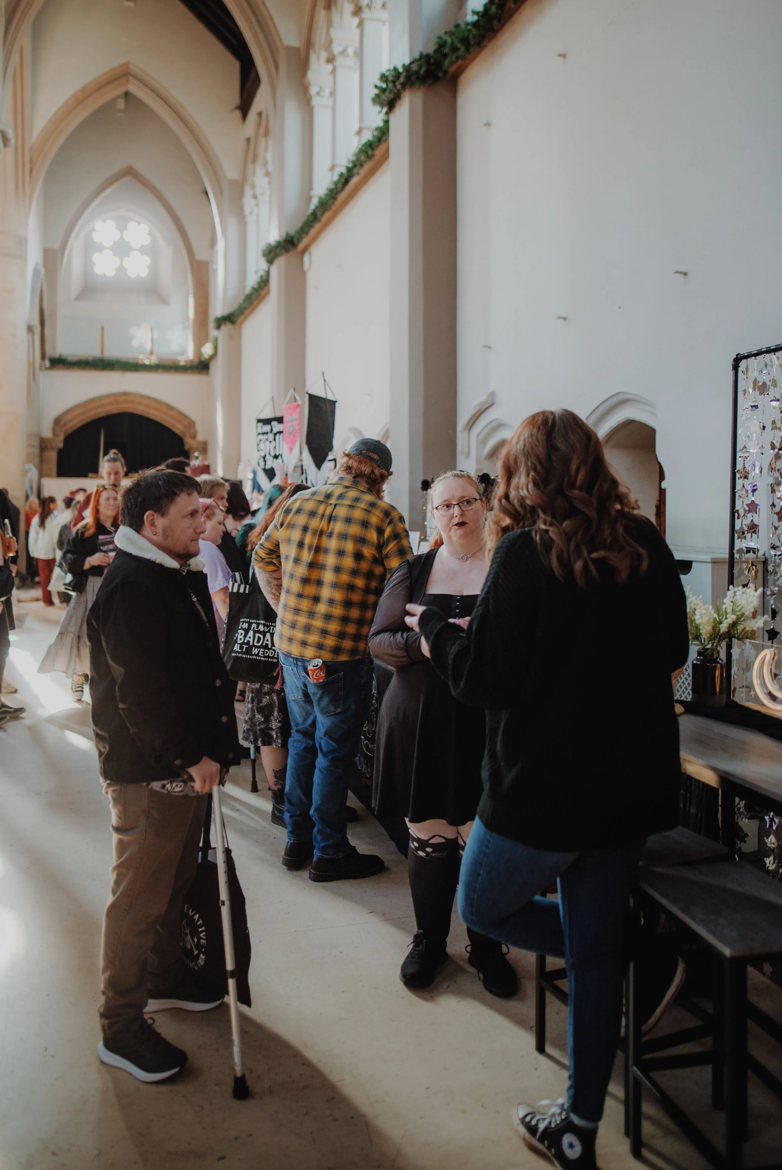 People standing and talking inside a large, arched, cathedral-like building with high ceilings, stained glass windows, and decorative greenery.