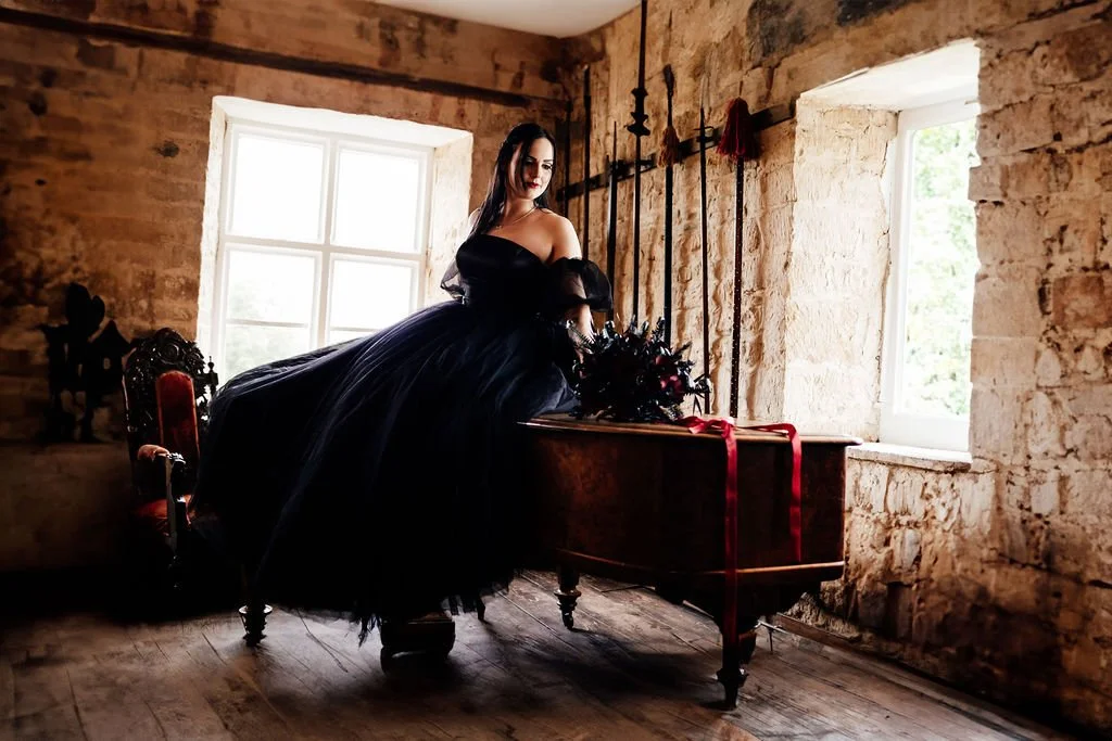 A woman in a black off-the-shoulder gown with a full tulle skirt stands in a rustic stone room, looking out a window. She has dark hair, red lipstick, and is beside a wooden table with a black floral arrangement and a red ribbon.