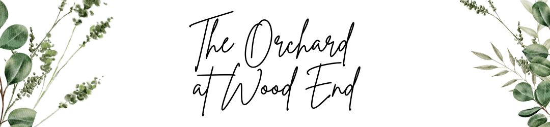 A white banner with green foliage on the left and right sides and the text 'The Orchard at Wood End' in black cursive font in the center.