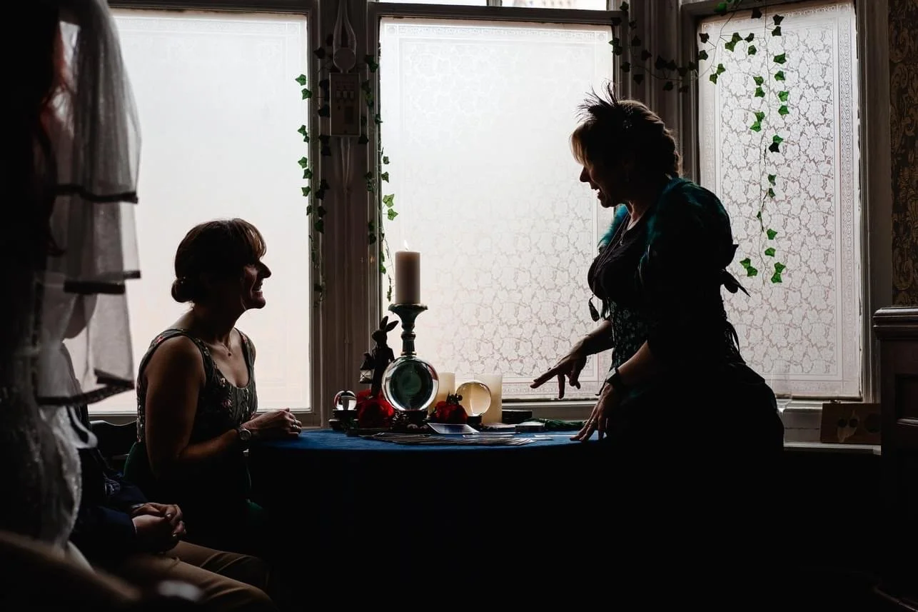 Silhouettes of two women in a room with large lace-covered windows. One woman stands and points, the other women sits at a table decorated with candles and tarot. The scene appears to be indoors with natural light filtering through the windows.