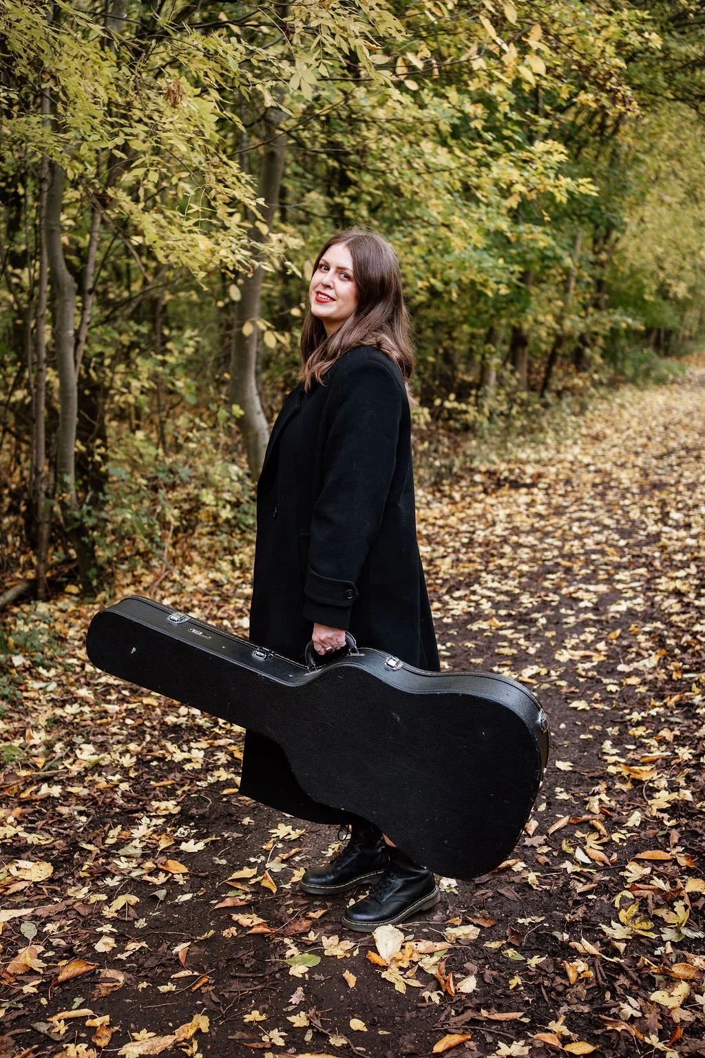A woman with brown hair, wearing a black coat and black boots, stands on a trail in autumn surrounded by yellow and green leaves, holding a black guitar case.