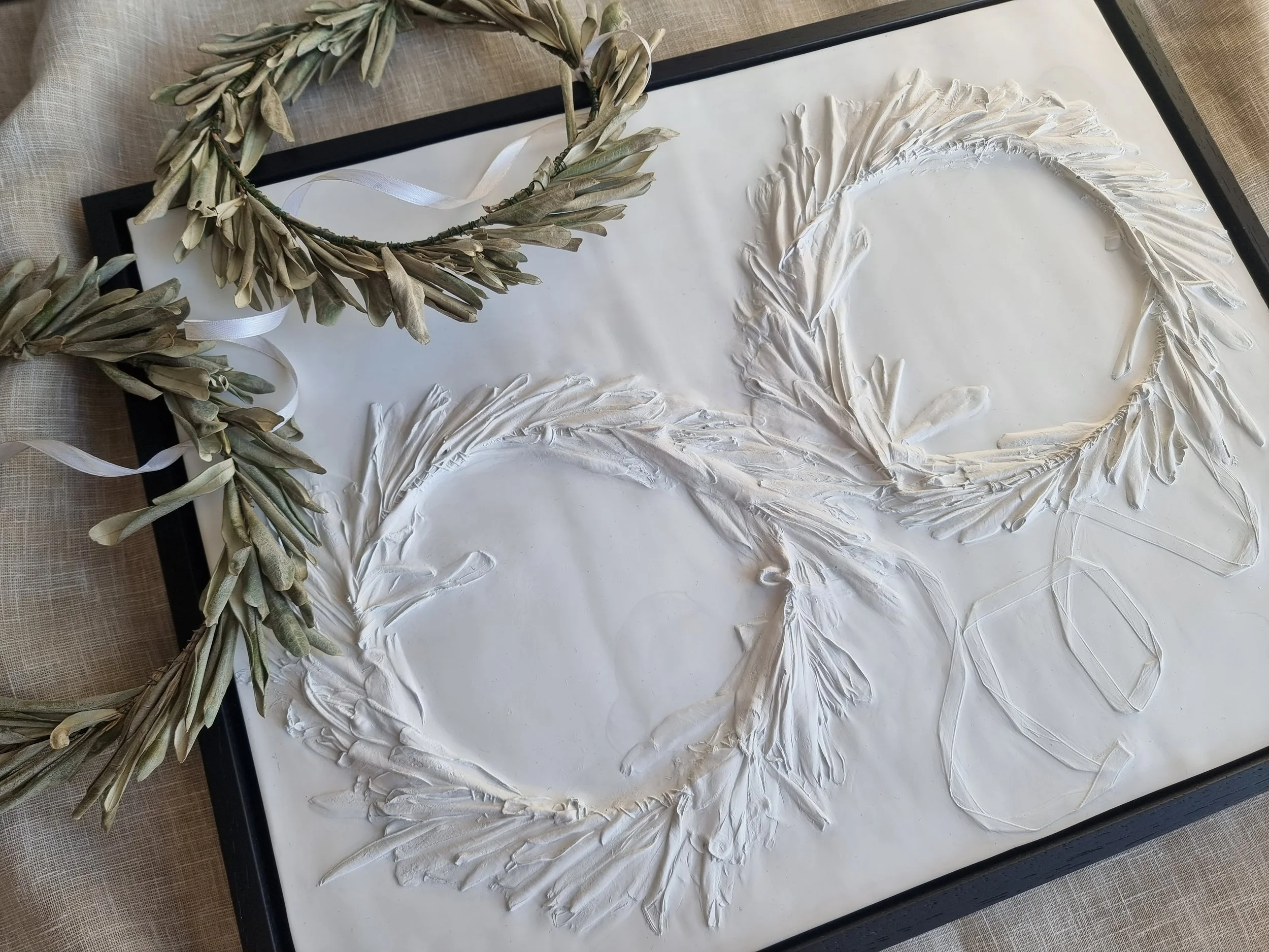 Two olive branch wreaths with white ribbons placed on a framed white textured art piece with embossed laurel and ribbon designs.
