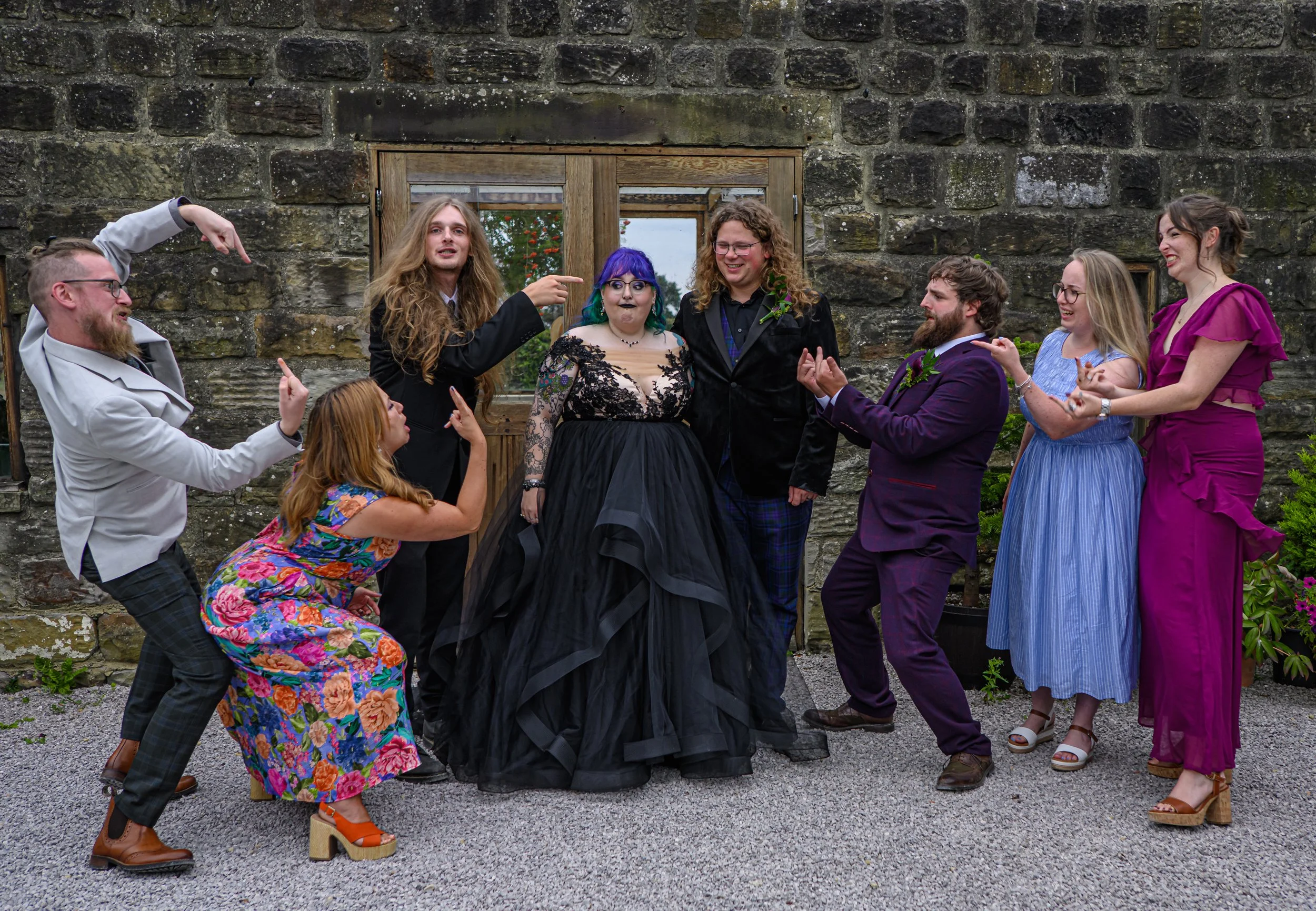 A group of nine diverse people are posing playfully in front of a stone wall and wooden window, smiling and making gestures. They are dressed in a variety of colorful, formal, and eccentric outfits, suggesting a festive or themed gathering.
