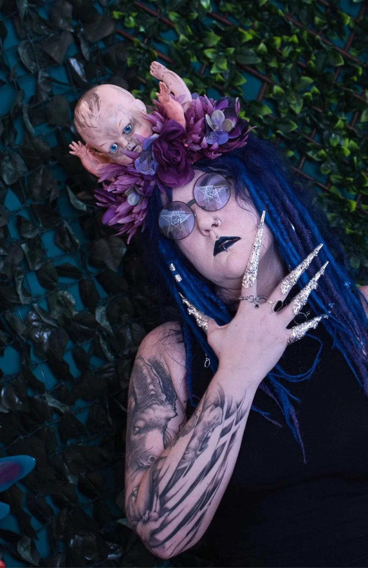 A person with blue and purple dreadlocks wearing reflective glasses, dark lipstick, and jewelry, lying against a leafy background. They have a headpiece featuring a doll's head surrounded by purple flowers and foliage.
