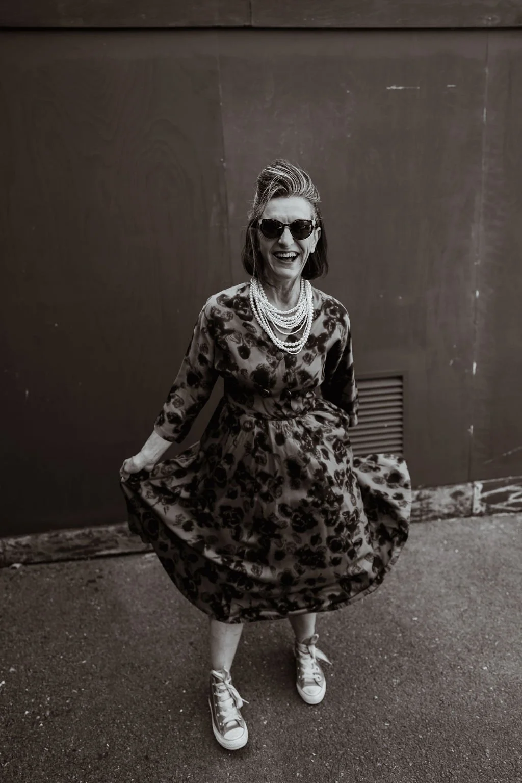 A woman wearing sunglasses, a floral dress, and multiple pearl necklaces, smiling and holding out the sides of her dress while standing on a sidewalk in front of a plain wall.