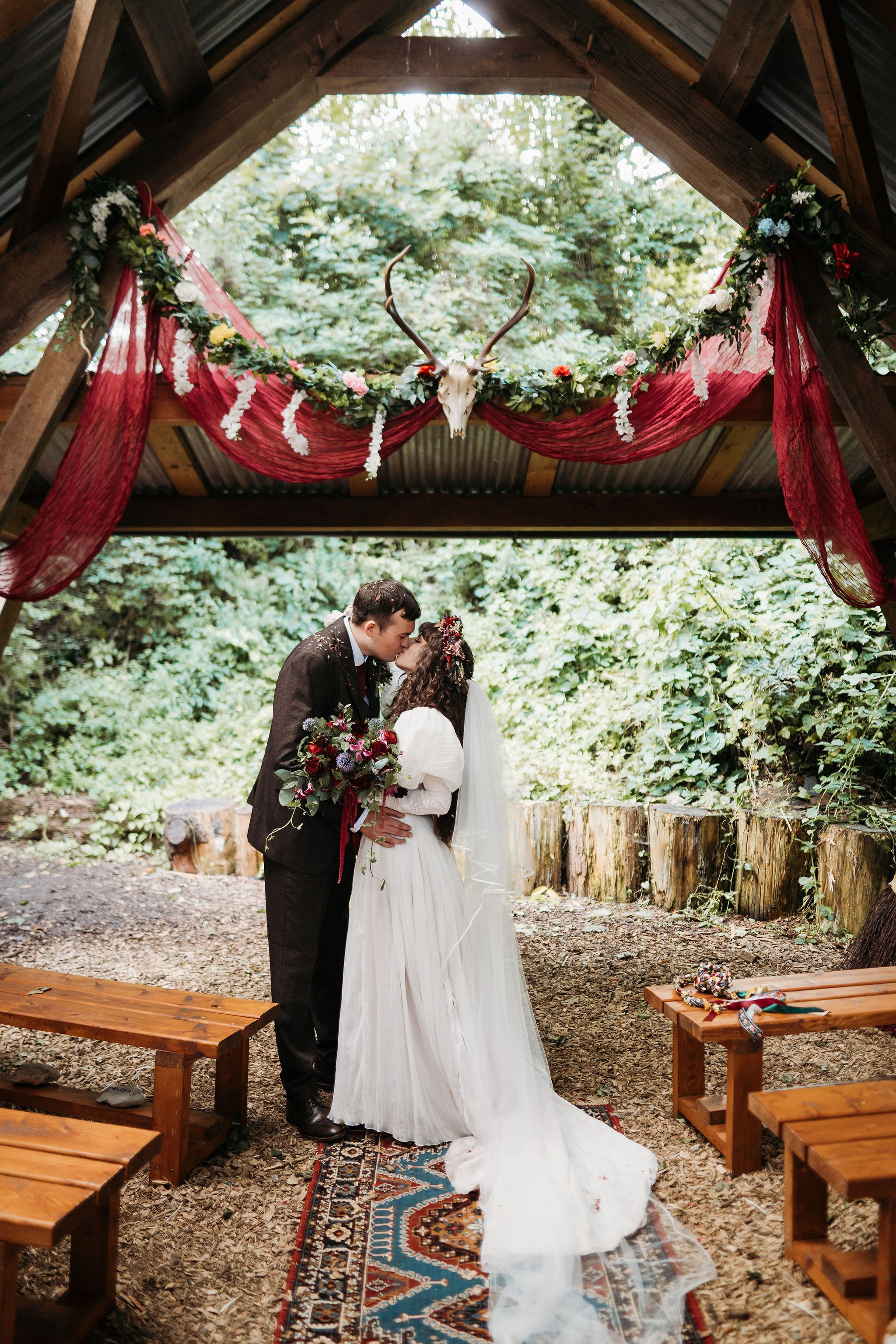 A couple in wedding attire sharing a kiss under a rustic wooden pavilion decorated with red fabric, white flowers, greenery, and a deer skull with antlers.