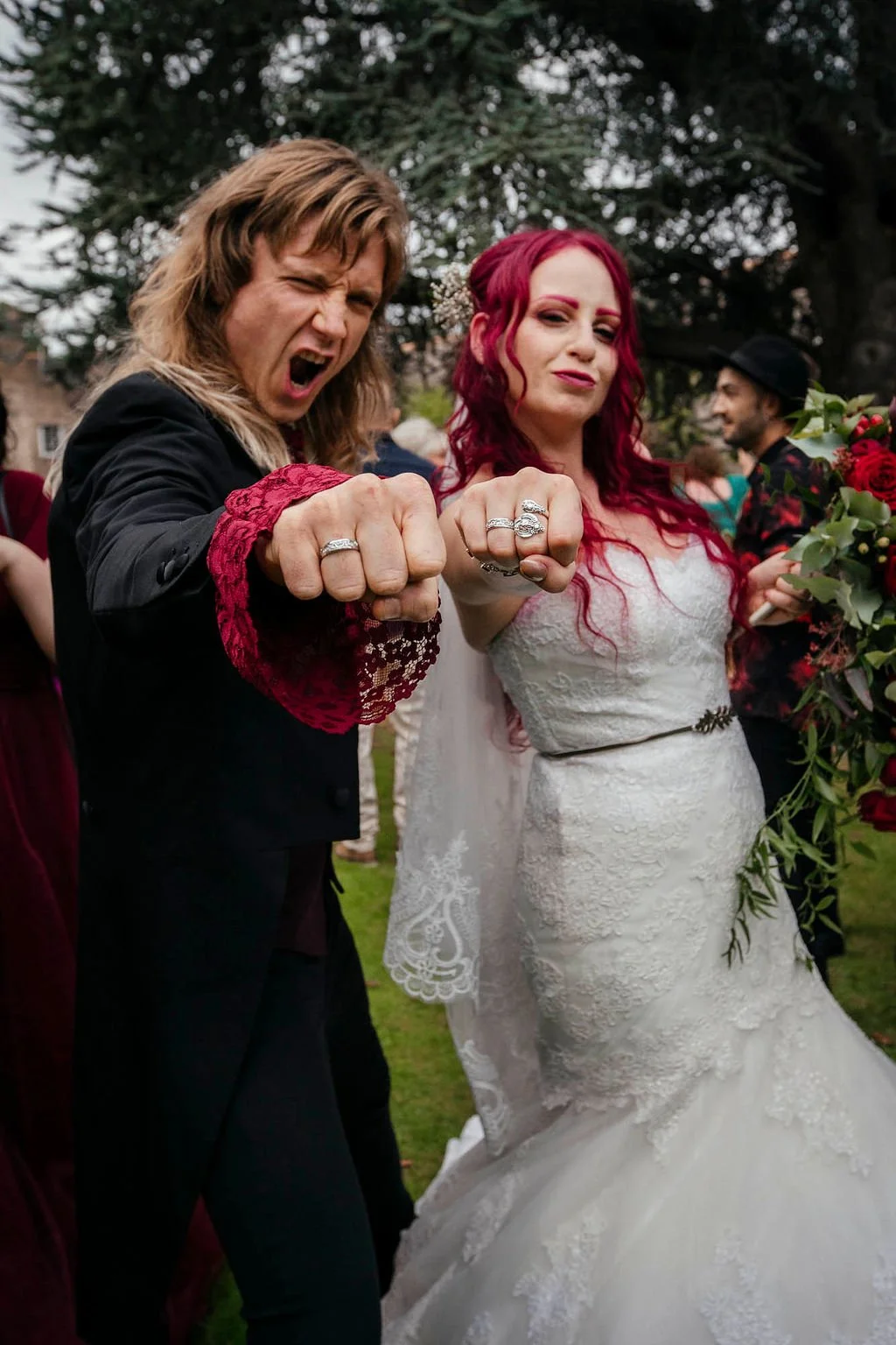 Two people, a man and a woman, dressed in wedding attire, showing off their wedding rings and making playful fists with their hands at a wedding celebration outdoors.
