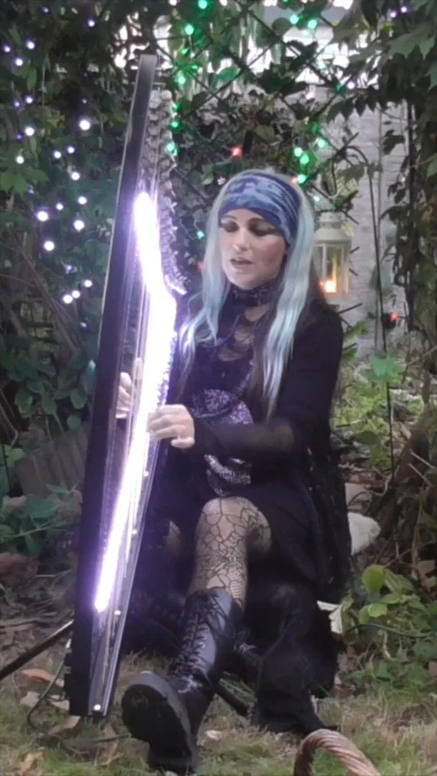 A woman with long, silver-blue hair, wearing a patterned headscarf, lace gloves, and black clothing, sitting outdoors surrounded by greenery and string lights, holding a long, illuminated object that resembles a sword or light fixture.