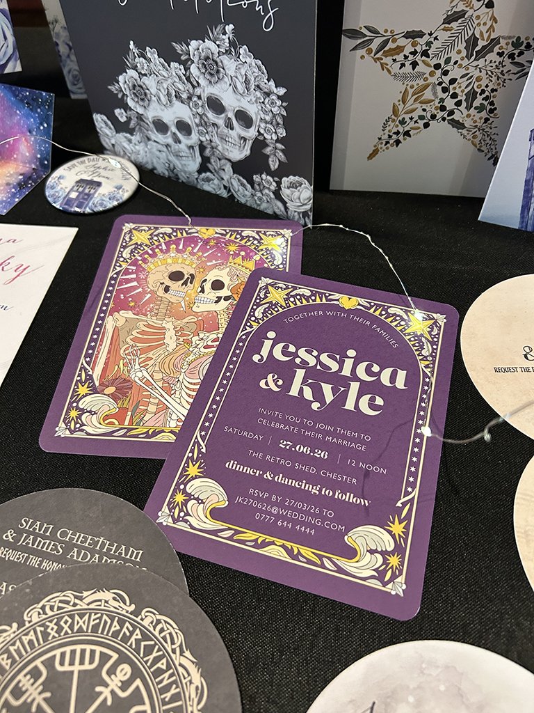 Purple wedding invitation for Jessica and Kyle with skeleton and star illustration, details of the event at The Retro Shed in Chester, plus nearby skeleton-themed cards on a black table.