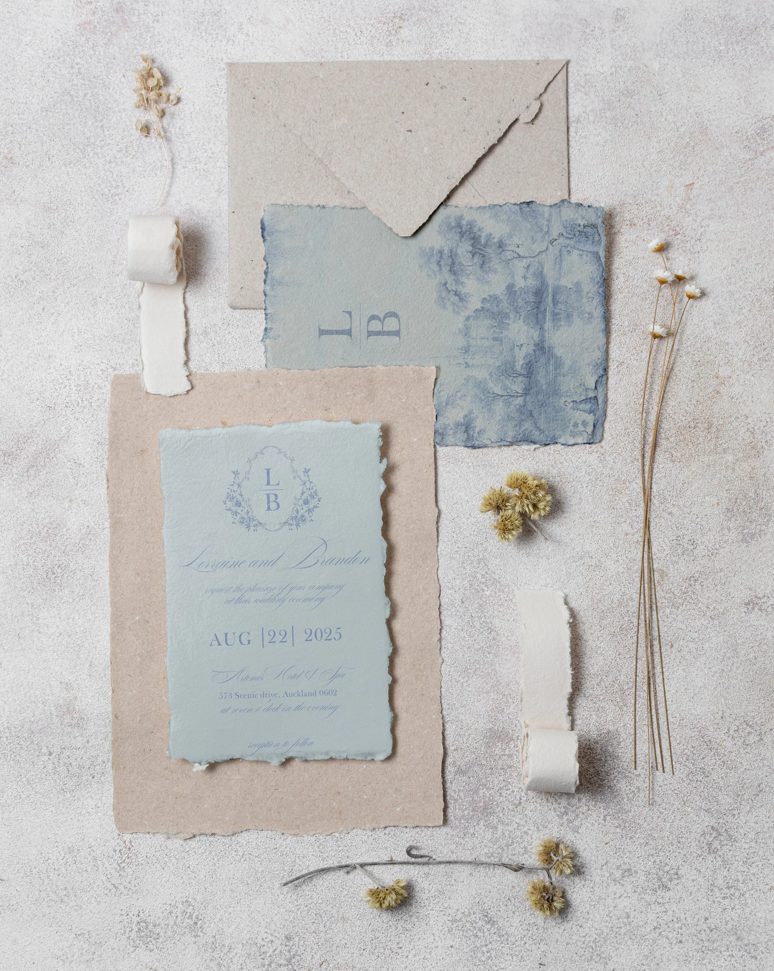 Wedding invitation suite with torn textured paper and dried flowers, featuring a light blue invitation card with dark blue text, placed on a neutral textured background.