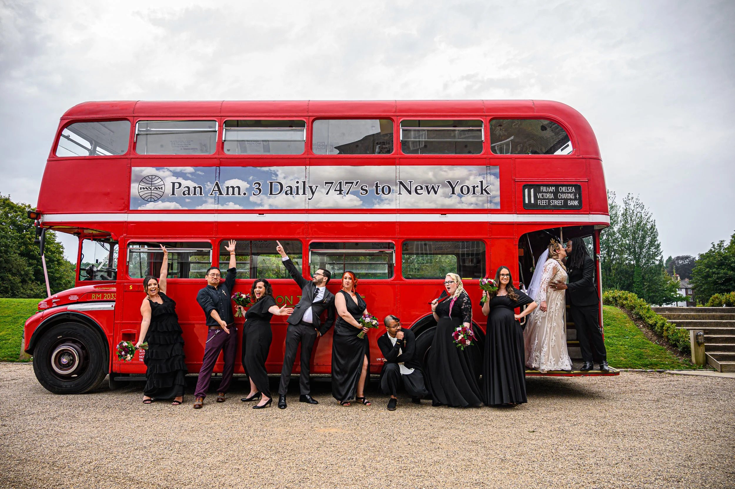 Group of wedding guests and a bride and groom standing in front of a red double-decker bus, with some guests cheering and others kissing, on a cloudy day in a park.