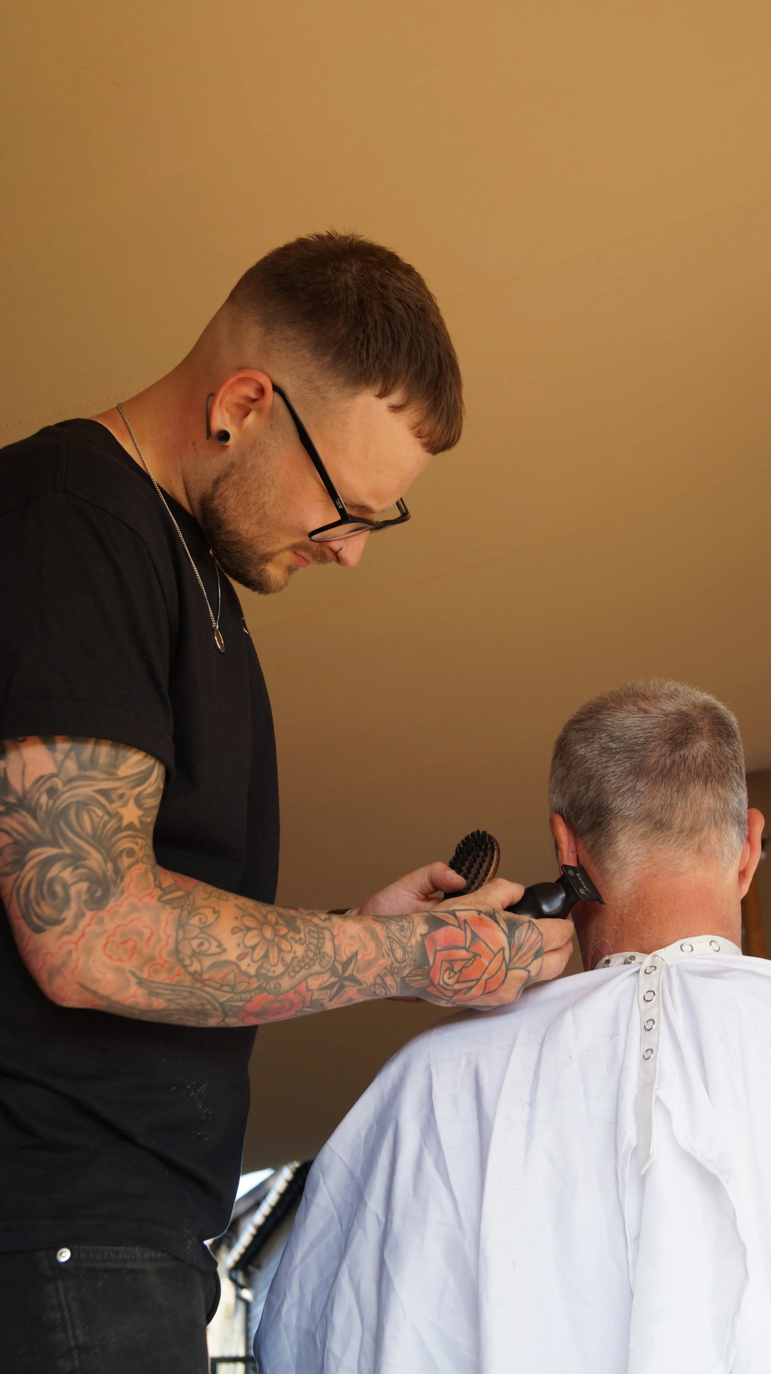 A tattooed hairstylist trimming a man's hair with clippers in a salon.