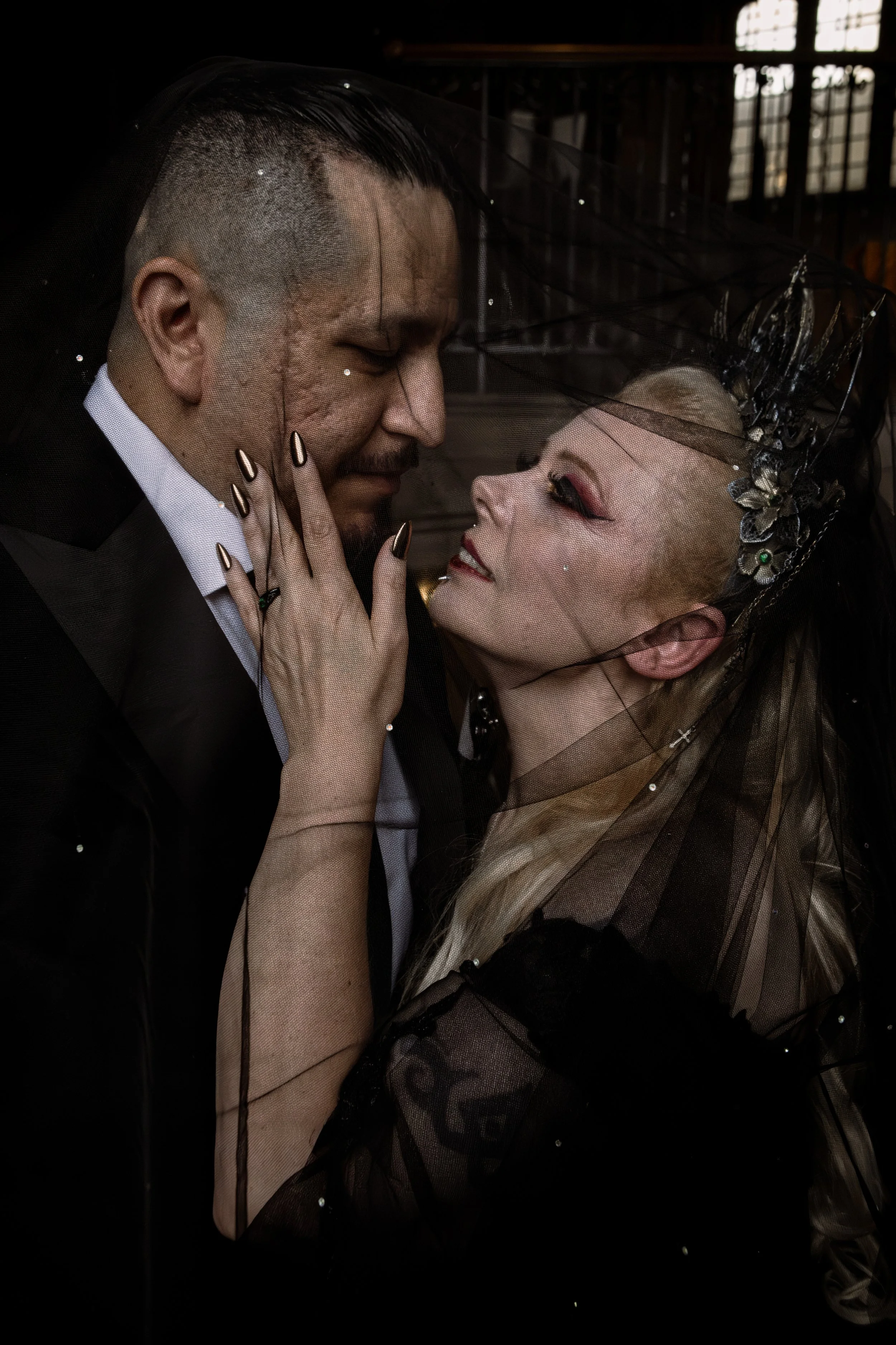 Gothic newlyweds embrace under the brides black veil, her hand gently holds her husbands face