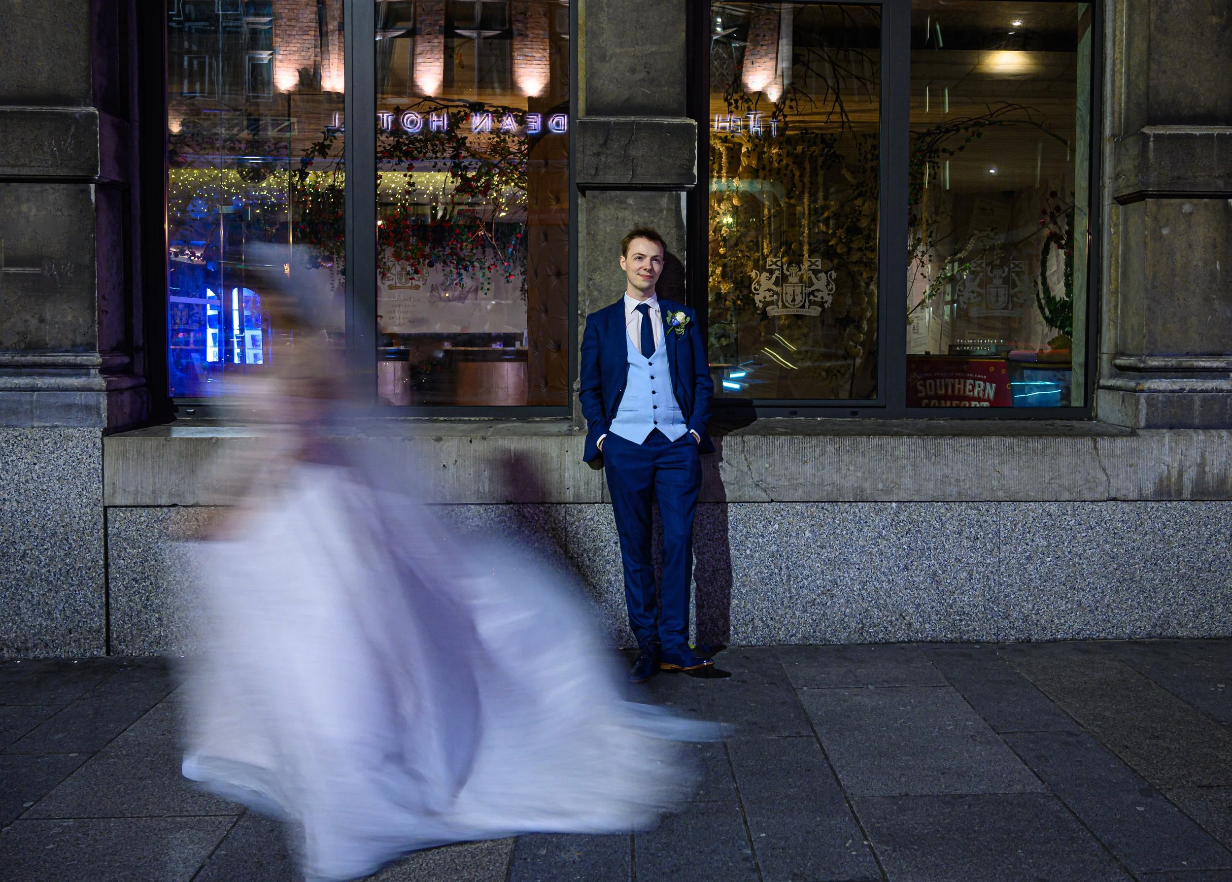 A man in a blue suit and white shirt leaning against a building wall at night, with a blurred figure in a white dress passing by in front of him.
