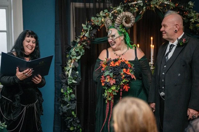 A woman reading from a script, a person dressed in a gothic costume holding a bouquet, and a man in a suit standing under a decorated arch with candles in the background.