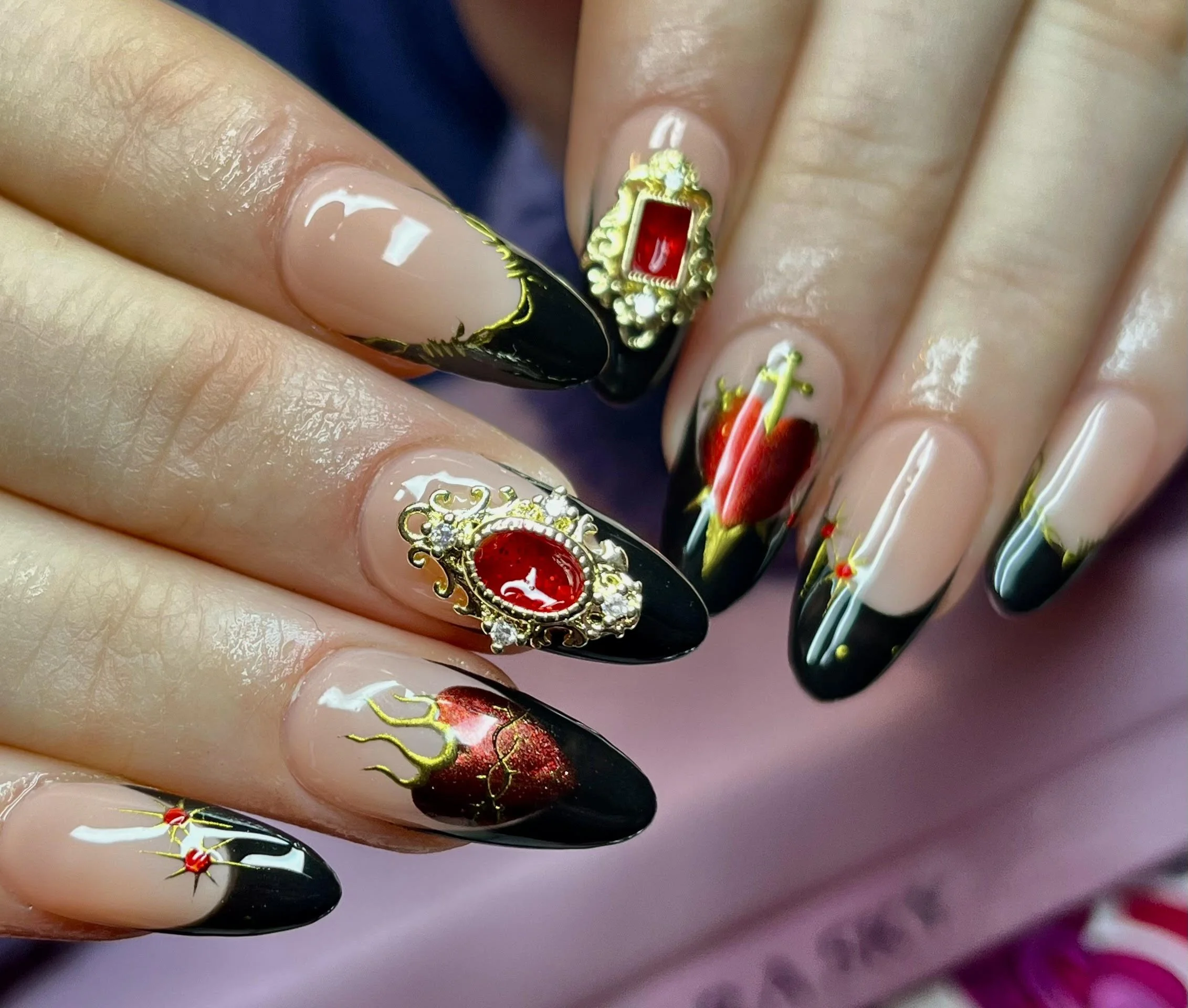 Close-up of manicured nails with elaborate red and black nail art, gold accents, and ornate jewelry embellishments.