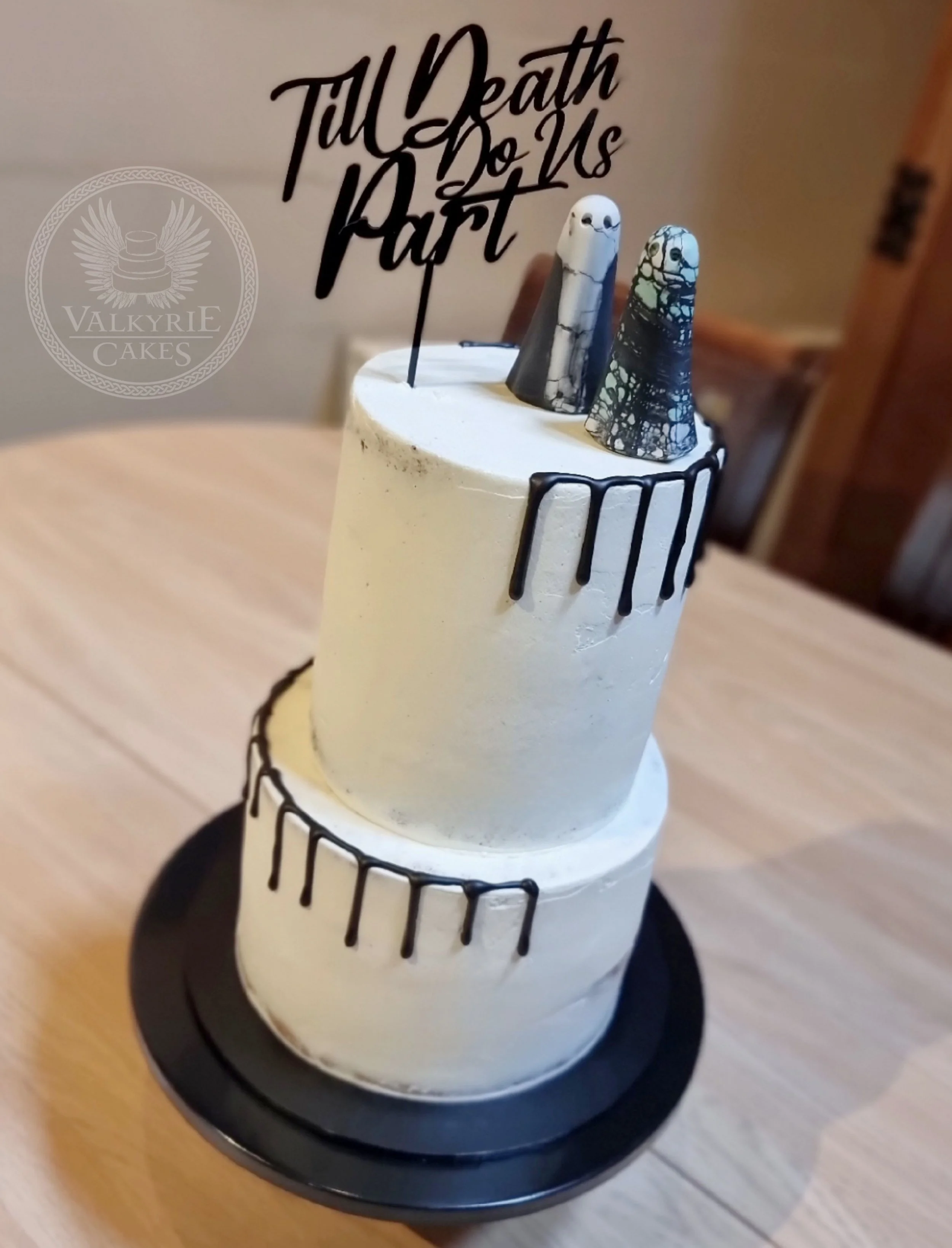A two-tiered cake with white frosting decorated with black chocolate drips, topped with two marbled fondant skulls, and a cake topper that says 'Till Death Do Us Part' in black script. The cake is placed on a black round cake stand.