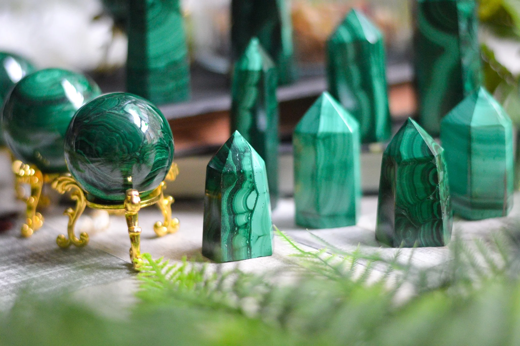 Collection of polished malachite stones in various shapes, including pyramids and spheres, displayed on a surface with some greenery and a blurred background.