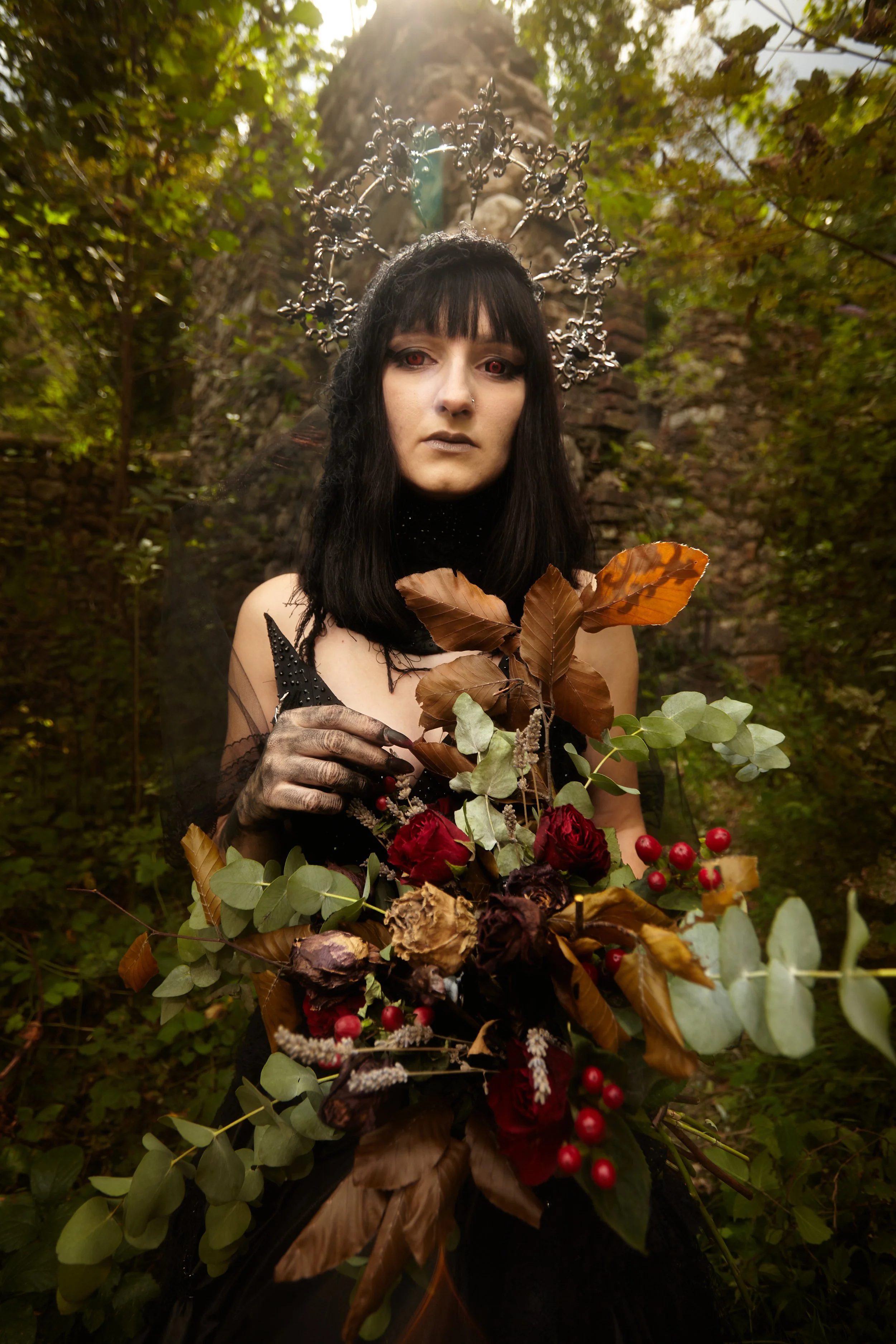 A woman with long black hair, wearing dark makeup and an elaborate metallic halo headpiece, stands outdoors in a wooded area, holding a large bouquet of dried and fresh leaves, flowers, and berries.