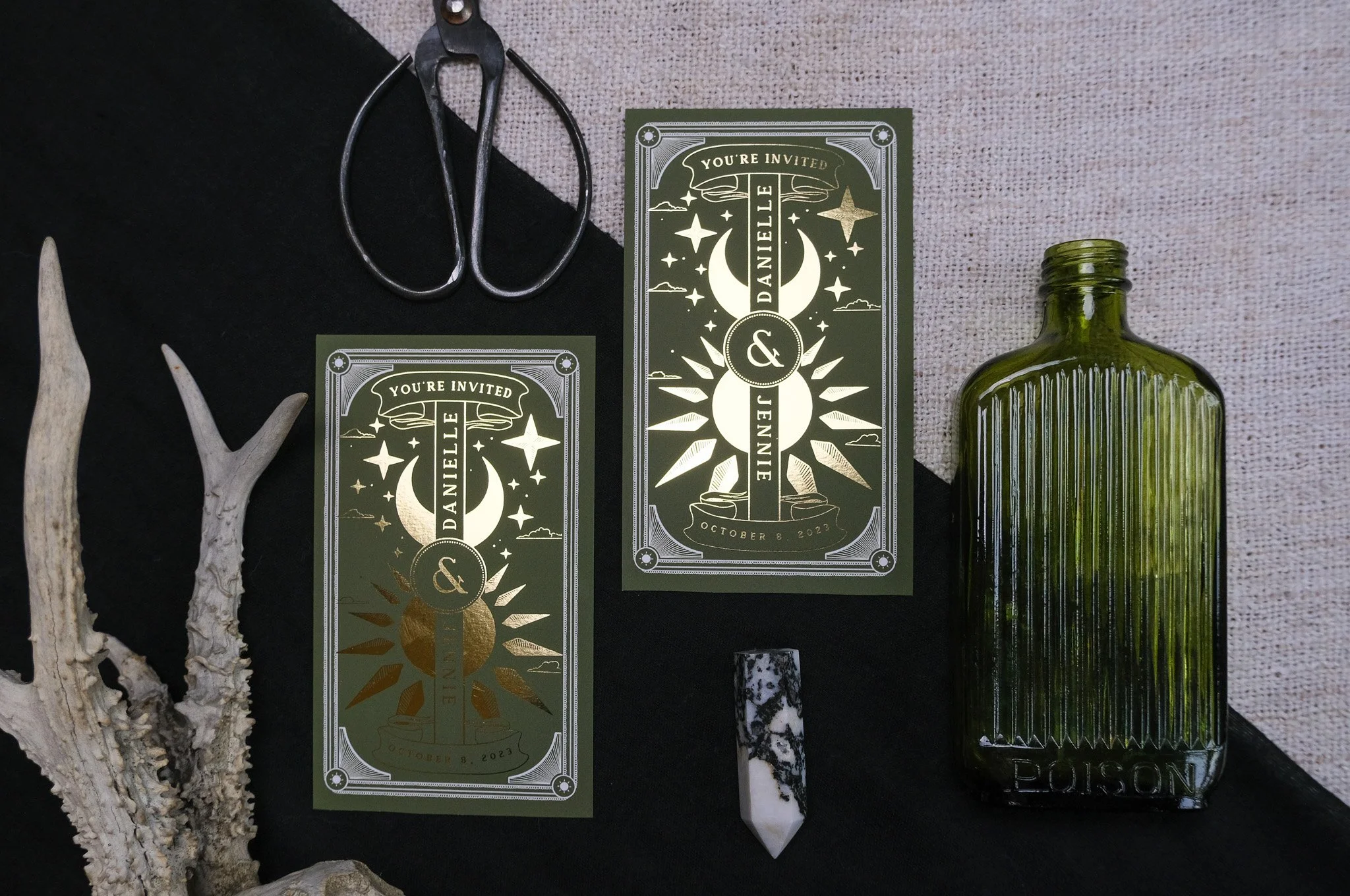 Dark green invitation cards with celestial designs, a black metal pair of scissors, a white and black marbled pendant, a green glass potion bottle, and white antlers arranged on a black and beige fabric background.
