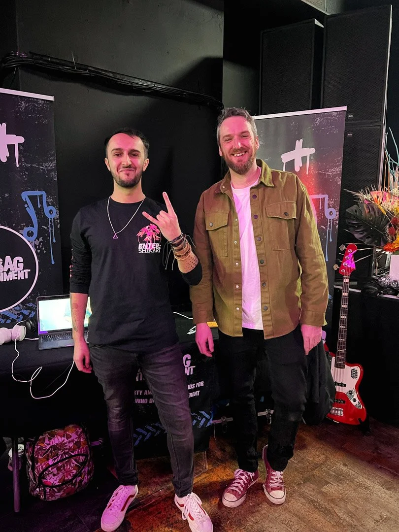 Two men standing next to each other smiling in front of music-themed banners, one wearing a black shirt and the other wearing a tan jacket, with a guitar and laptop visible in the background.