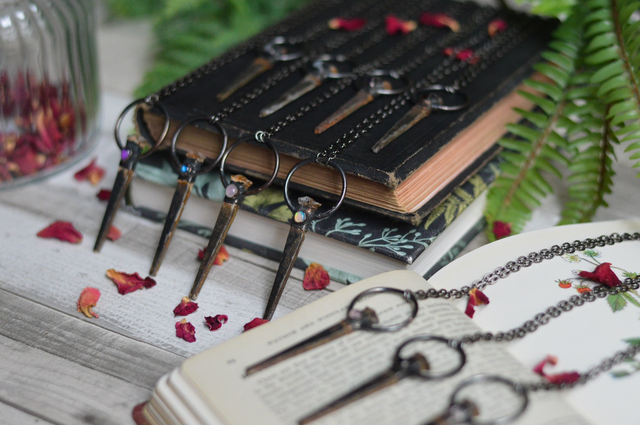 An assortment of antique keys with keychains placed on an open book, with more keys on a stack of books and dried flower petals scattered around, along with a fern plant in the background.