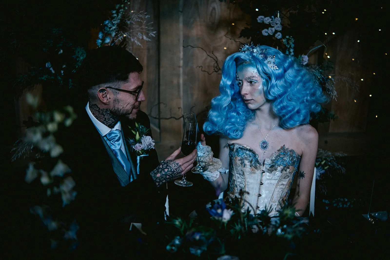 A man and a woman with blue hair sitting at a table with dark wood background and floral decorations, engaging in a toast with wine glasses.