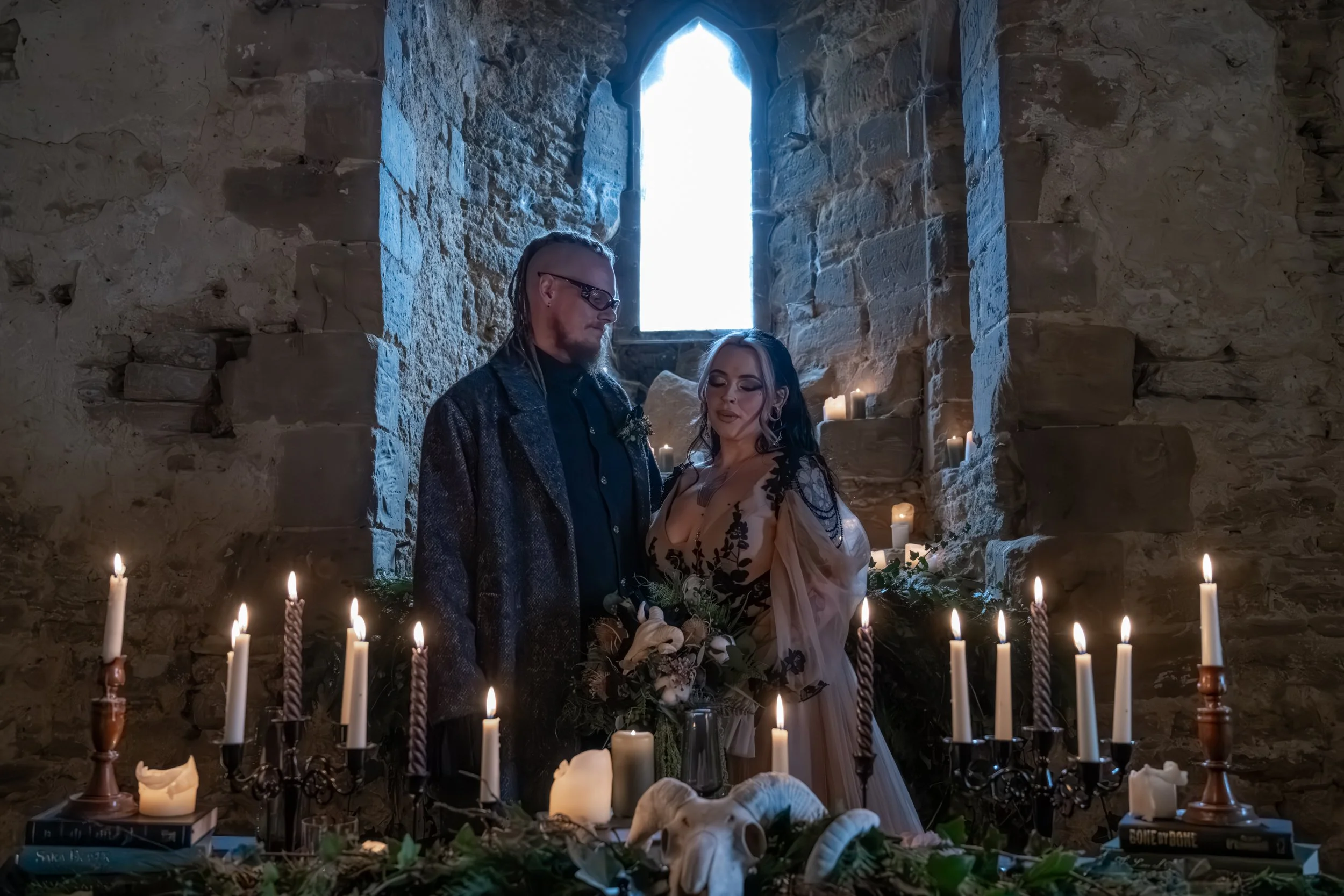 A couple stands inside a stone chapel, surrounded by lit candles and gothic decor, with a window casting light behind them, appearing to be in an intimate ceremony or photoshoot.