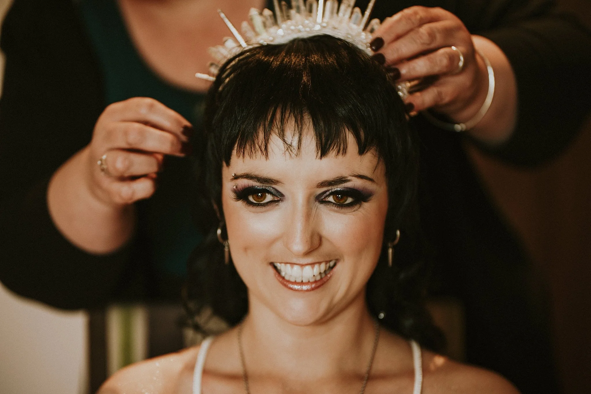 A woman with short black hair and brown eyes getting her hair adorned with a silver tiara by another person.