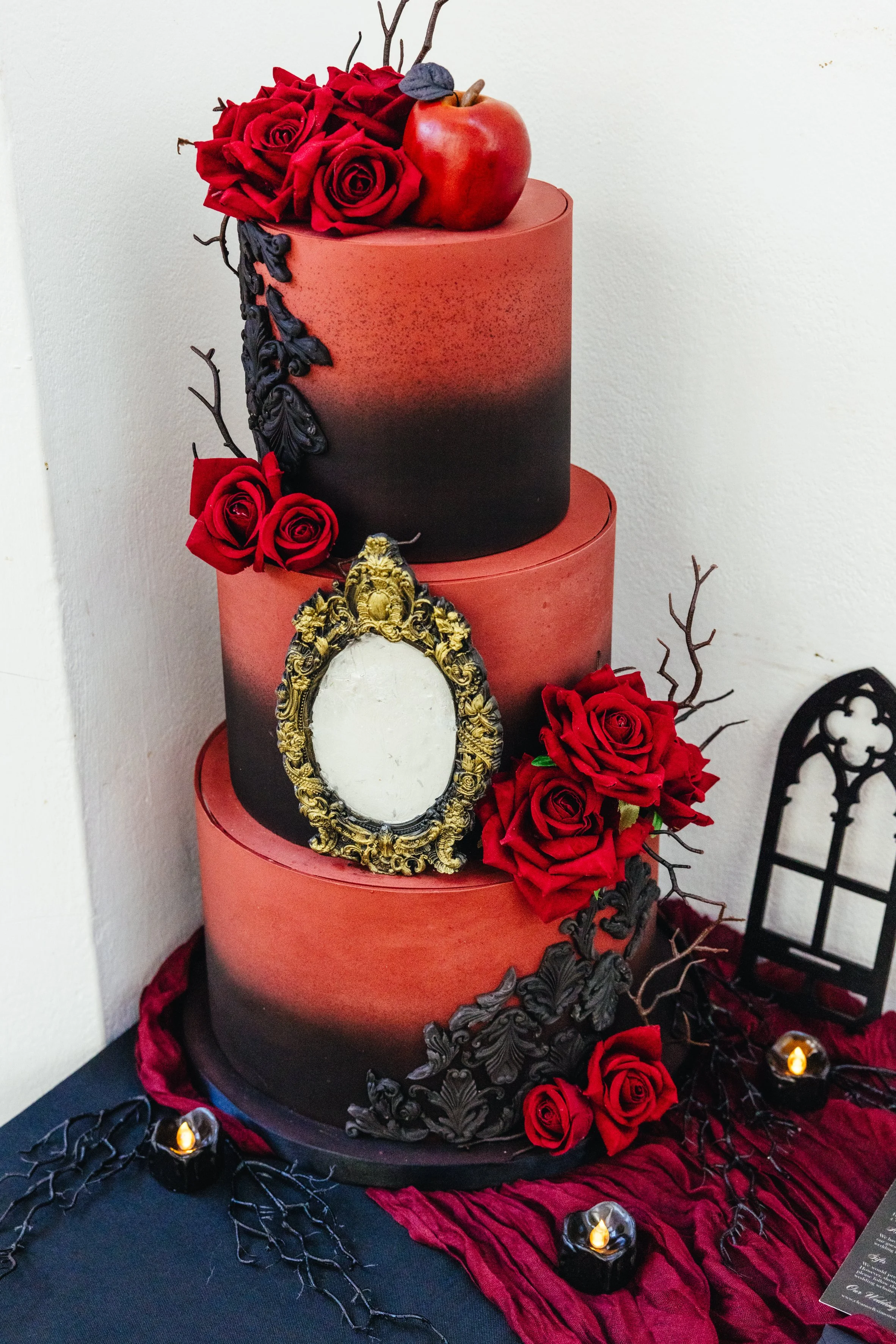 A three-tiered wedding cake with red and black gradient icing, decorated with red roses, black ornamental accents, and healthy green leaves. An apple and a small gray leaf are on top. A vintage gold and white frame with no photo stands on the middle 