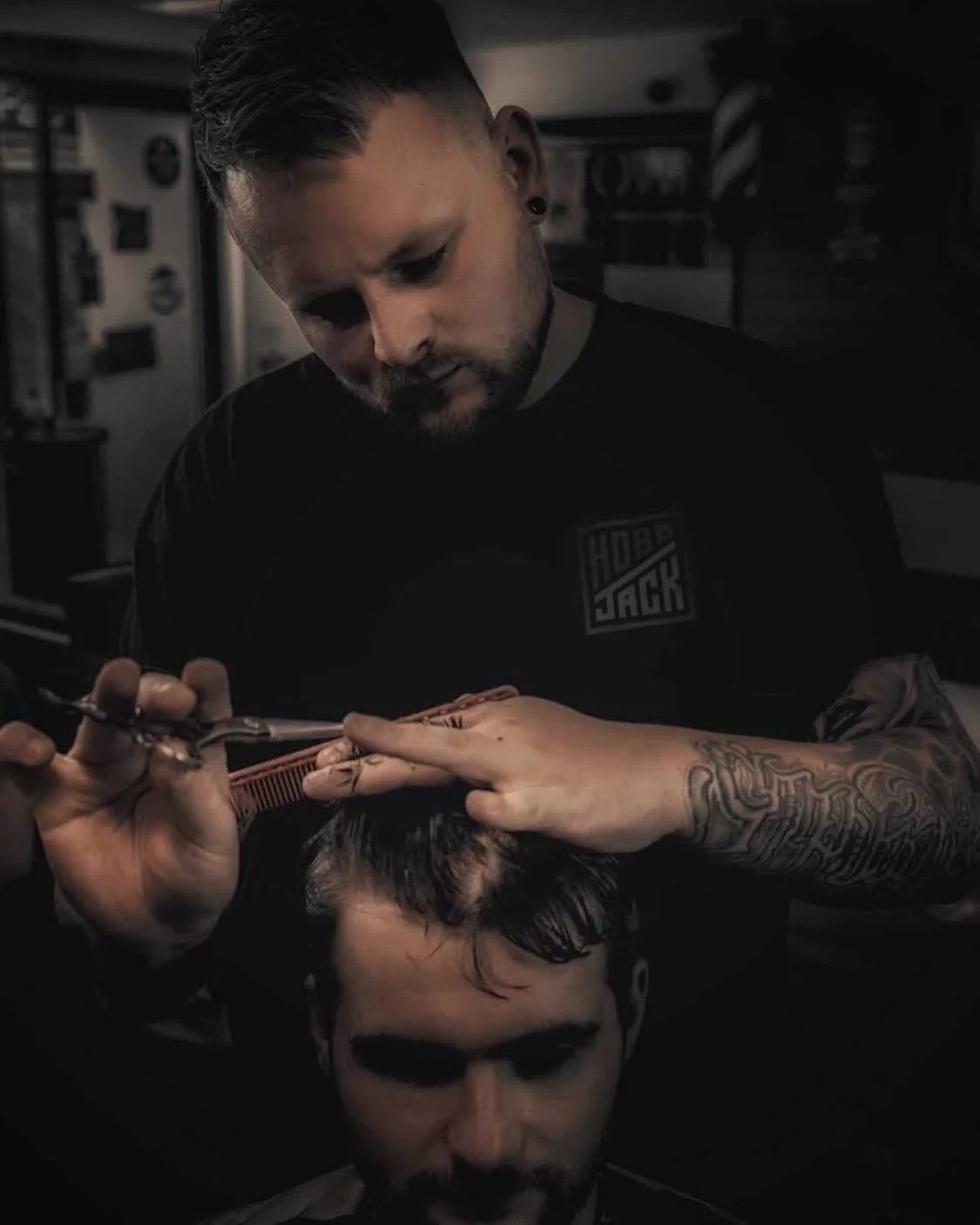 A barber with tattoos and earrings cuts a customer's dark hair with scissors in a barbershop.
