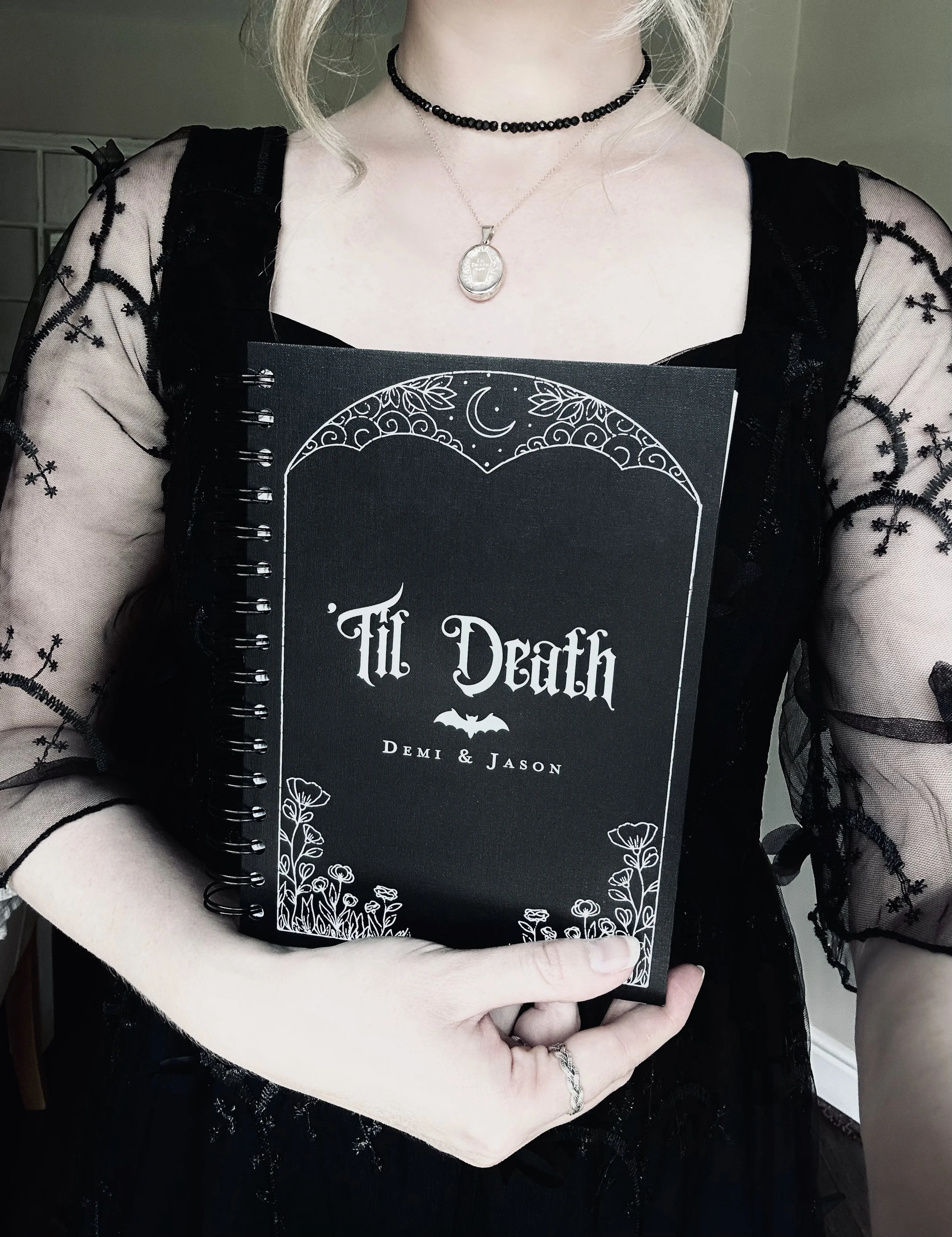 Person holding a notebook titled 'Til Death' with floral and moon illustrations, wearing a black lace dress, choker necklace, and a pendant necklace.