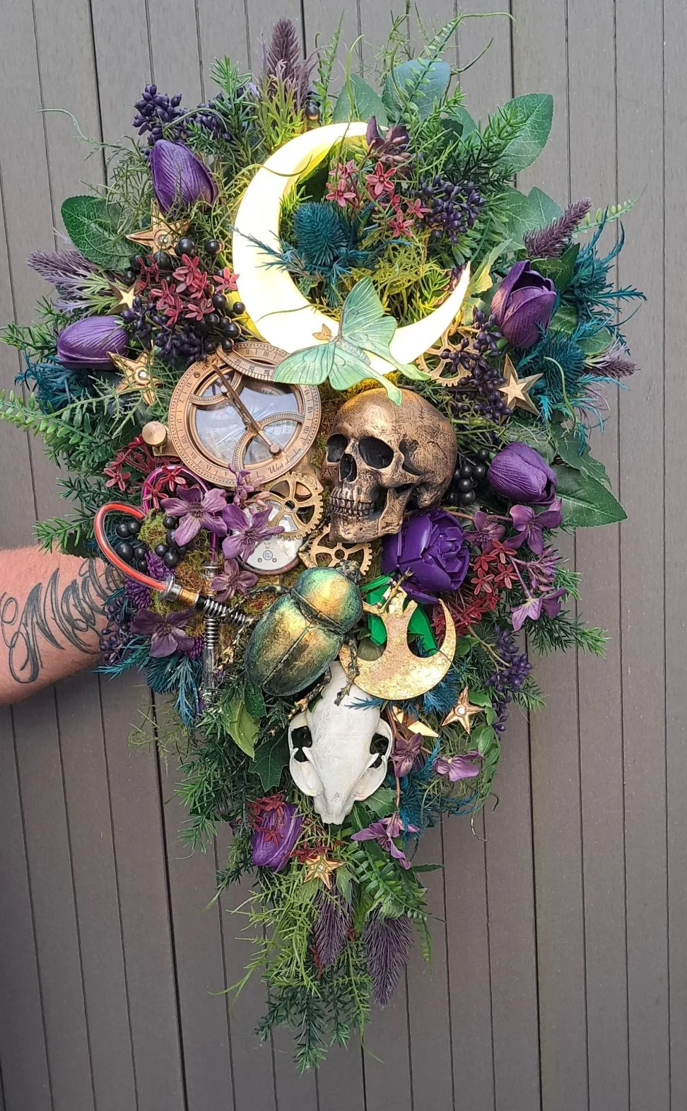 A decorative wall hanging with purple flowers, green foliage, a glowing crescent moon, a skull, a large beetle, a butterfly, and various other mystical and natural elements.