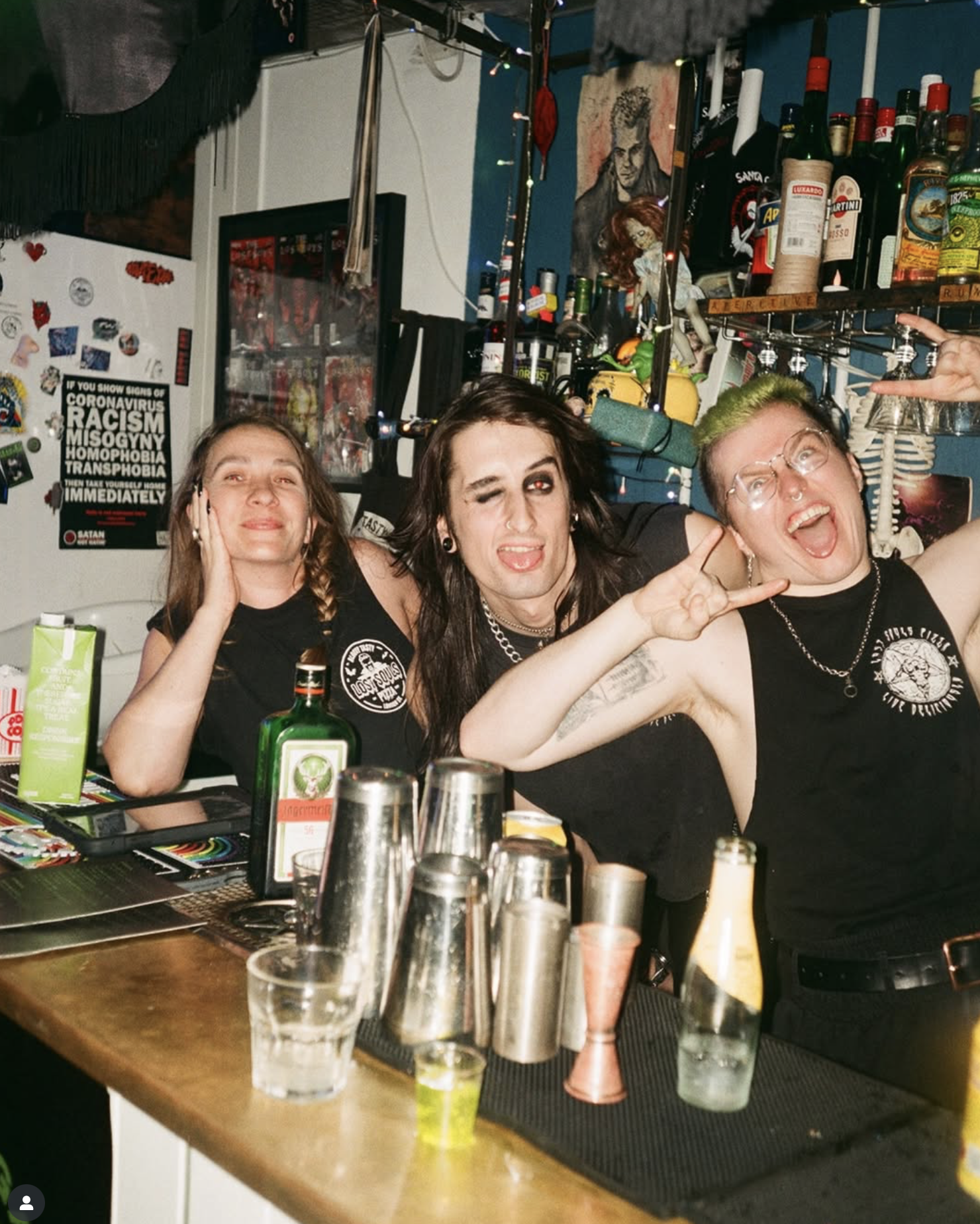 Three people at a bar, making playful expressions. The person on the left has long hair, the middle person has dark hair and is winking, and the person on the right has short green hair and is shouting. The bar counter has various bottles, glasses, a