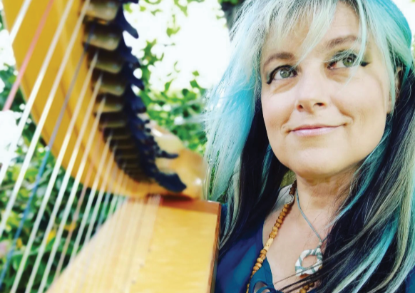 A woman with multi-colored blue, black, and blonde hair outdoors, smiling next to a large harp with green foliage in the background.