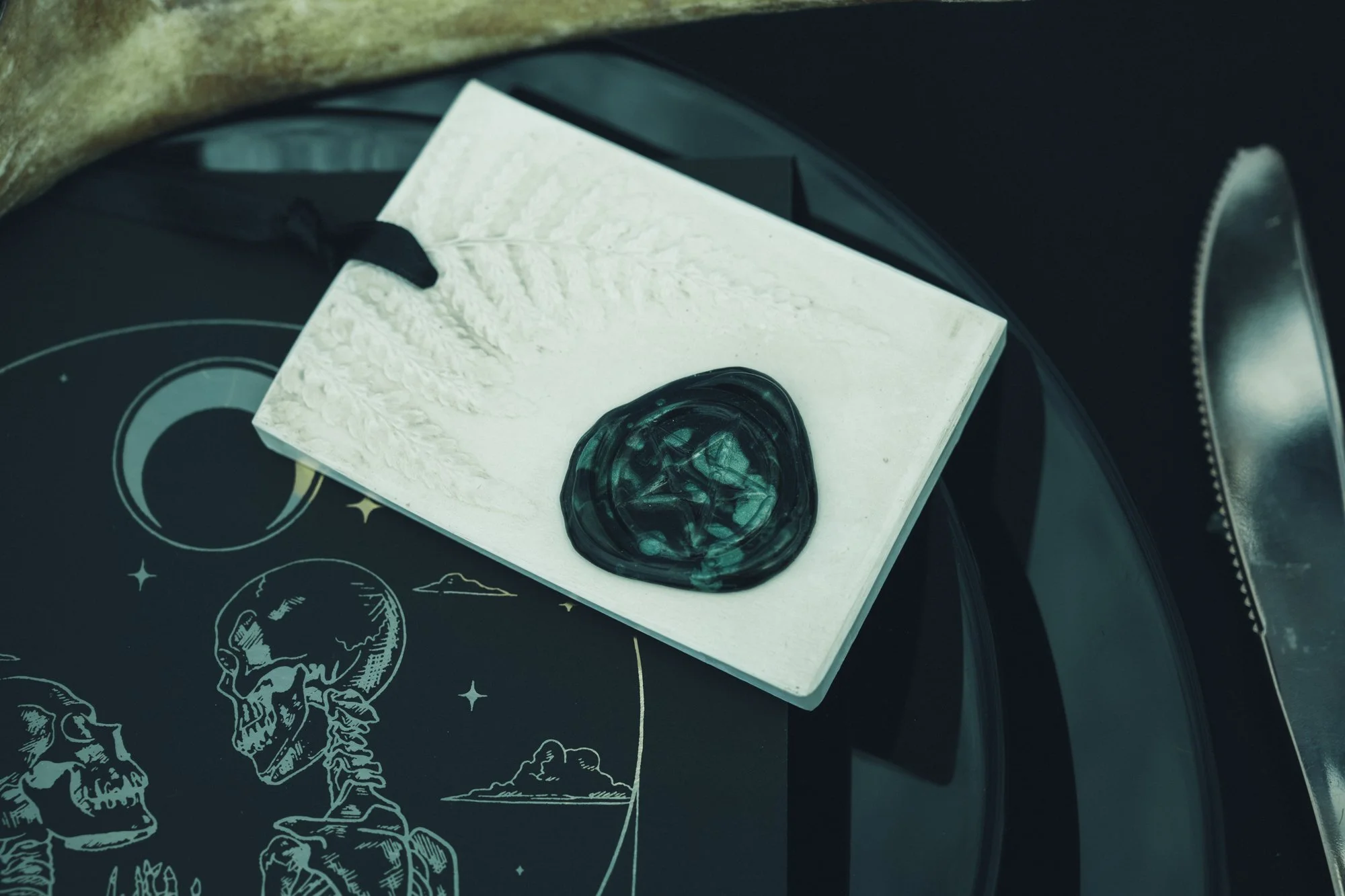 A white square card with a black wax seal rests on a black surface featuring illustrations of skulls and planets, with a glass lid of a record player nearby.