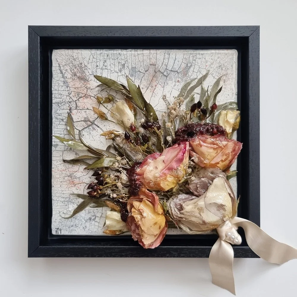 Pressed dried flowers and leaves framed in a black shadow box with a decorative ribbon on the bottom right corner.
