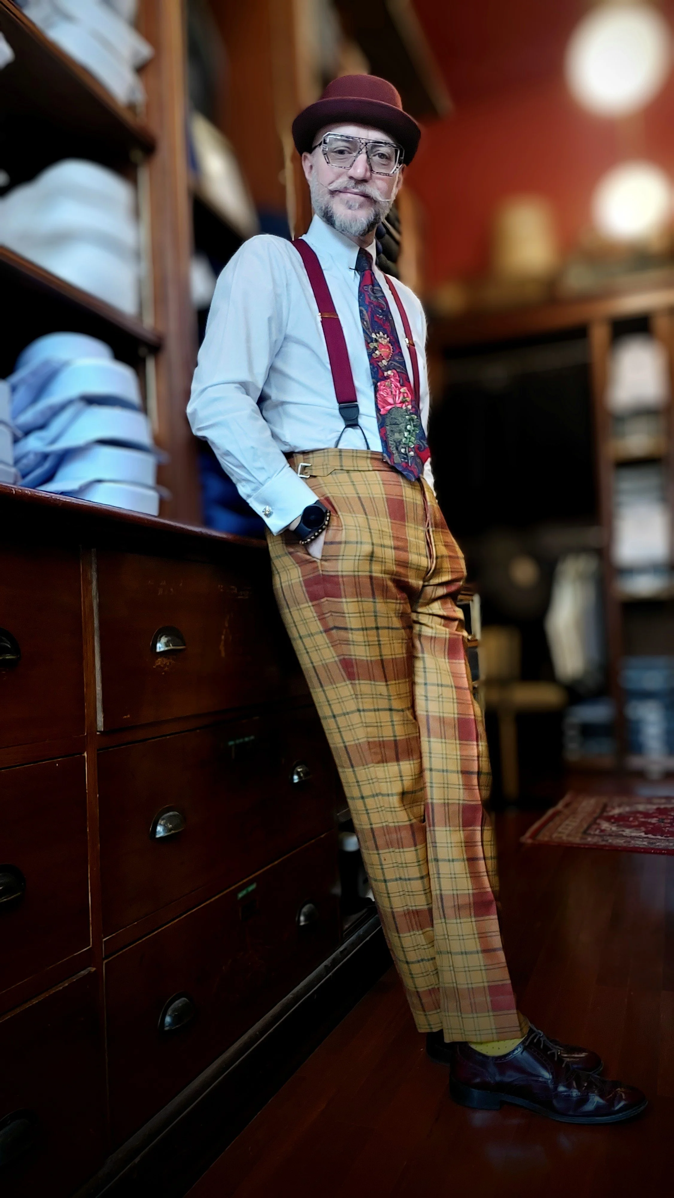 A man in colorful, patterned clothing stands in a vintage store with wooden shelves, wearing a hat, glasses, and a colorful dress shirt with suspenders. He has his hands in his pockets, leaning casually against a wooden dresser.