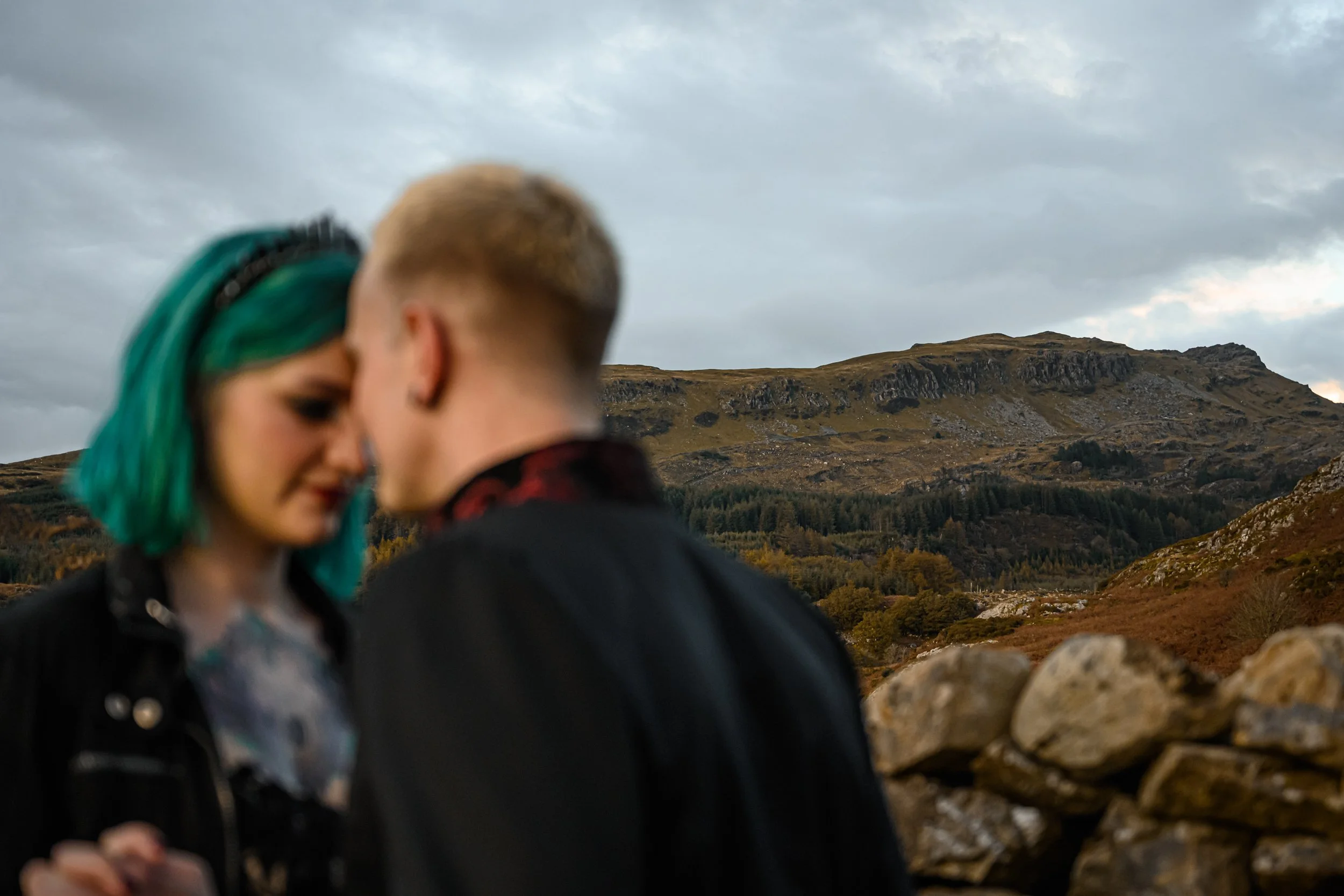 A couple with their foreheads touching outdoors with mountains and cloudy sky in the background.