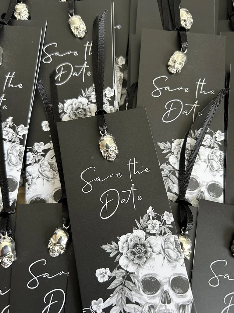 Black 'Save the Date' cards with white floral designs and skull embellishments, tied with black ribbons.