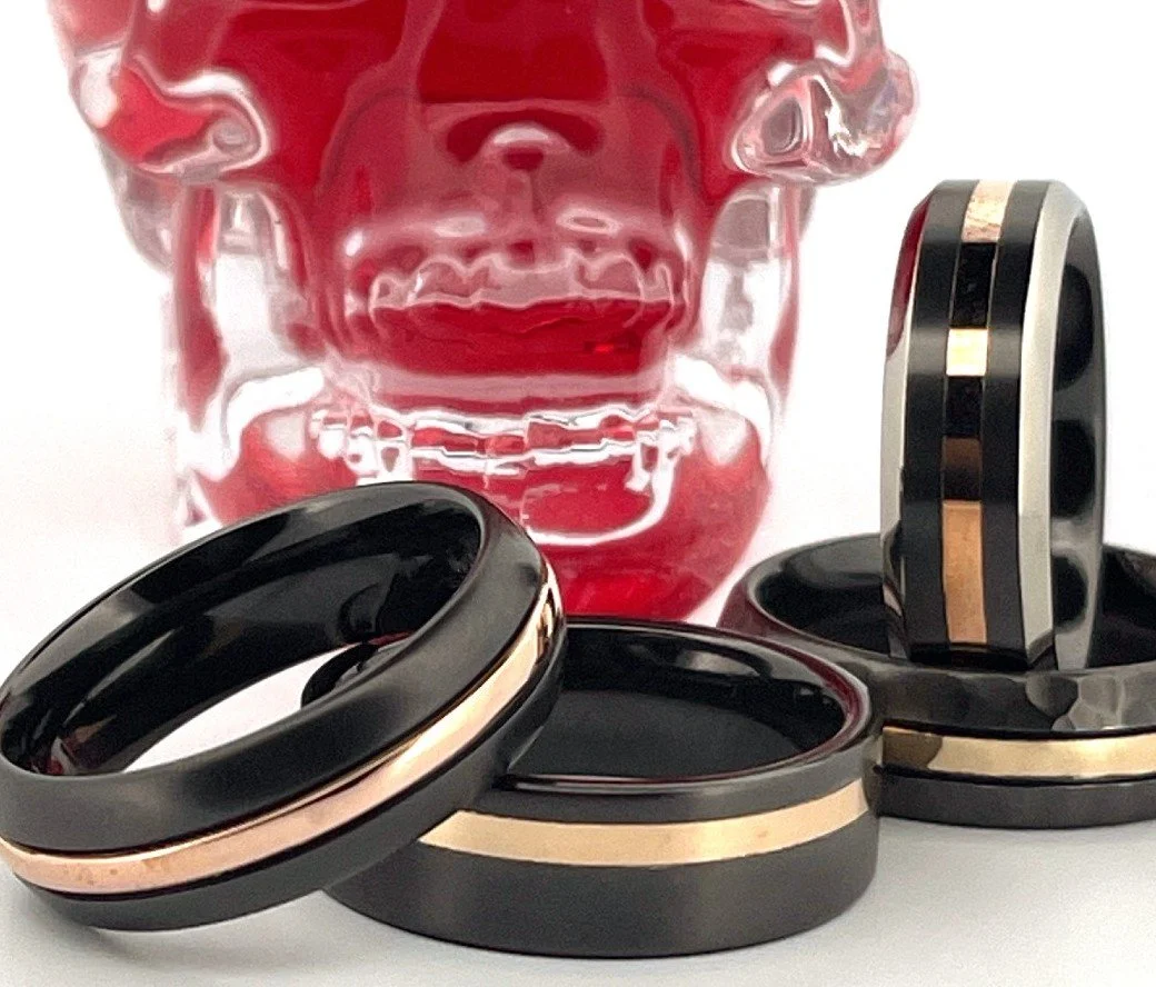 A clear glass skull sculpture with red accents behind black and metallic rings, including two rings with a gold band, arranged in front.
